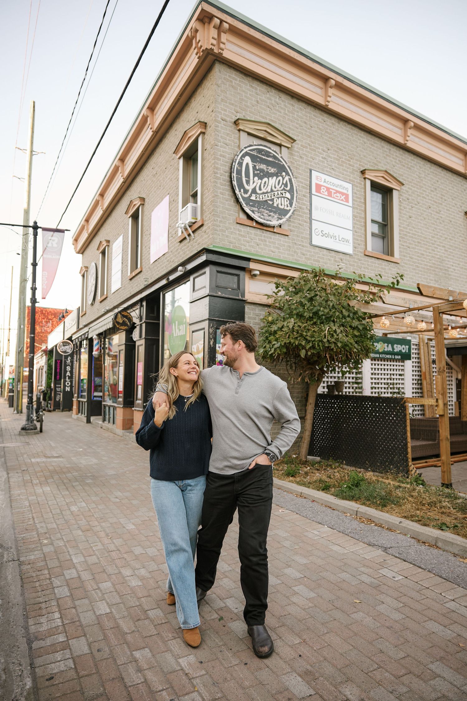 A couple walking and laughing together on a brick sidewalk outside a corner building.