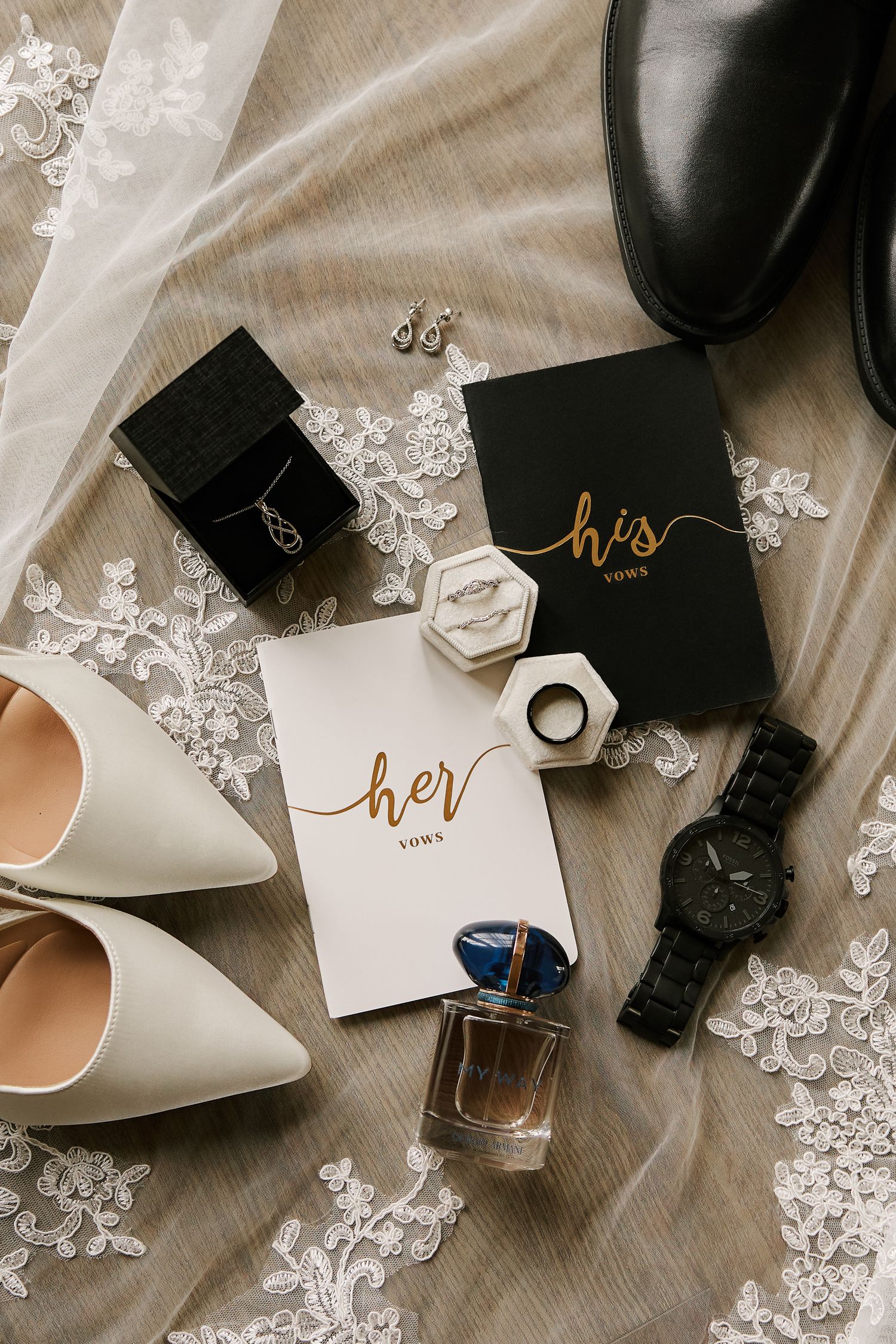 Wedding accessories including his and hers cards, shoes, and jewelry arranged on white fabric.