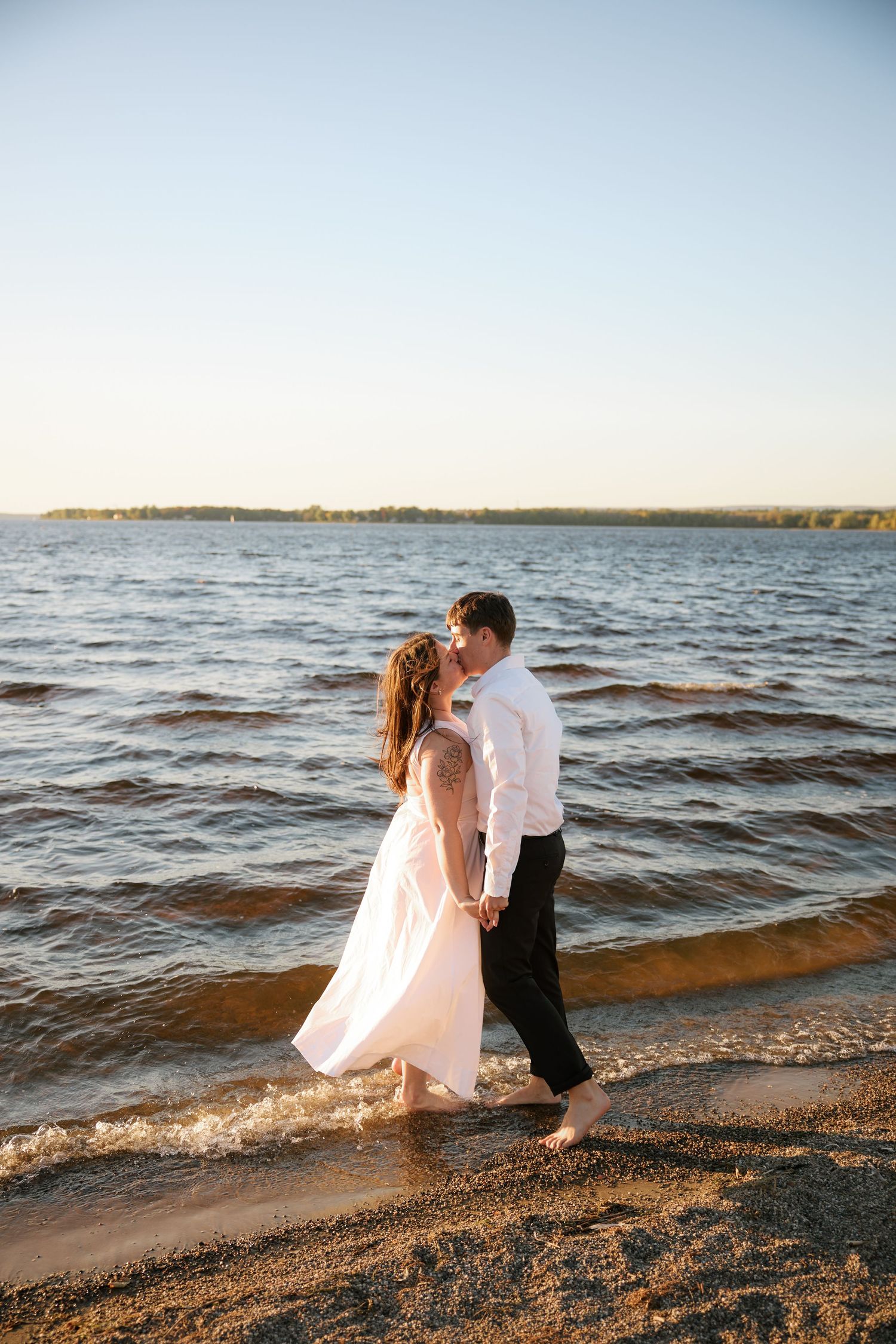 Couple sharing a romantic kiss on a sandy beach shore at golden hour, bride in white dress and groom in white shirt.