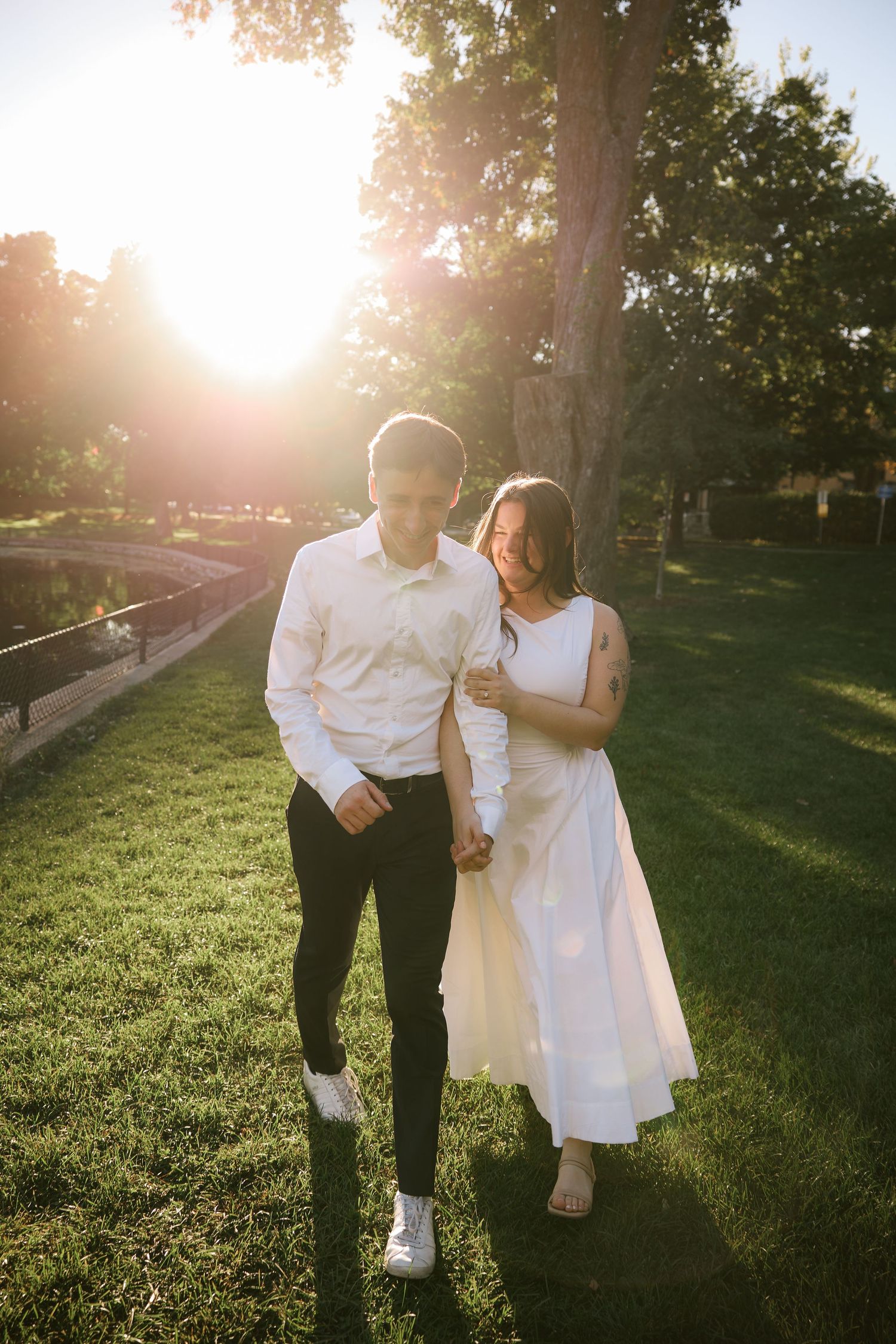Couple holding hands walking on green grass in a sunlit park, man in white shirt and black pants, woman in white dress.
