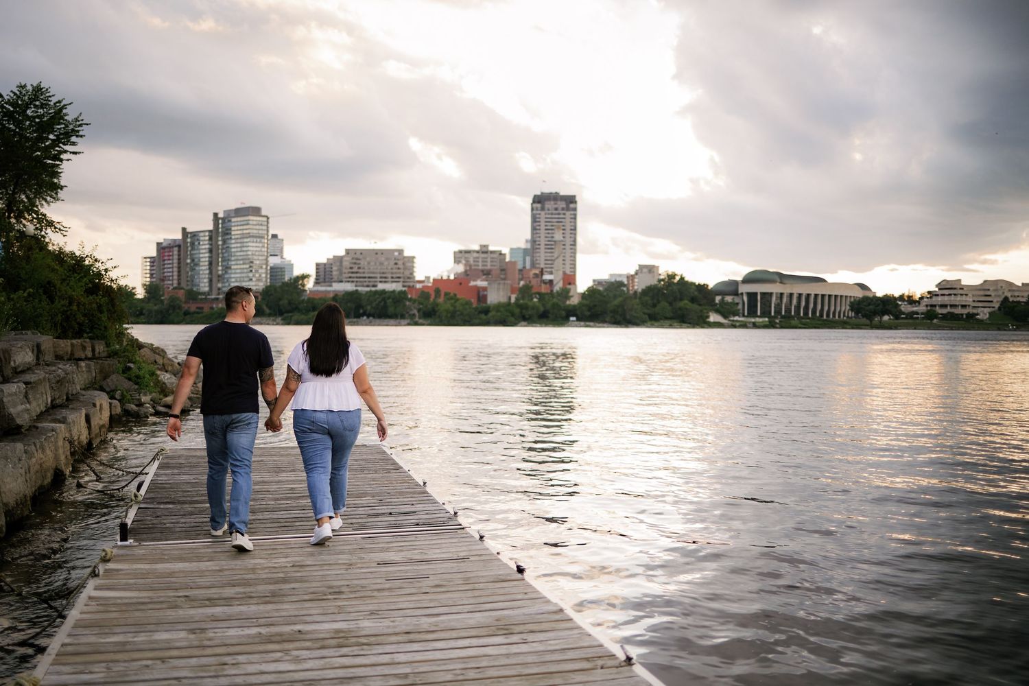 Couple holding hands walking on a wooden dock overlooking a city skyline and calm river at sunset.