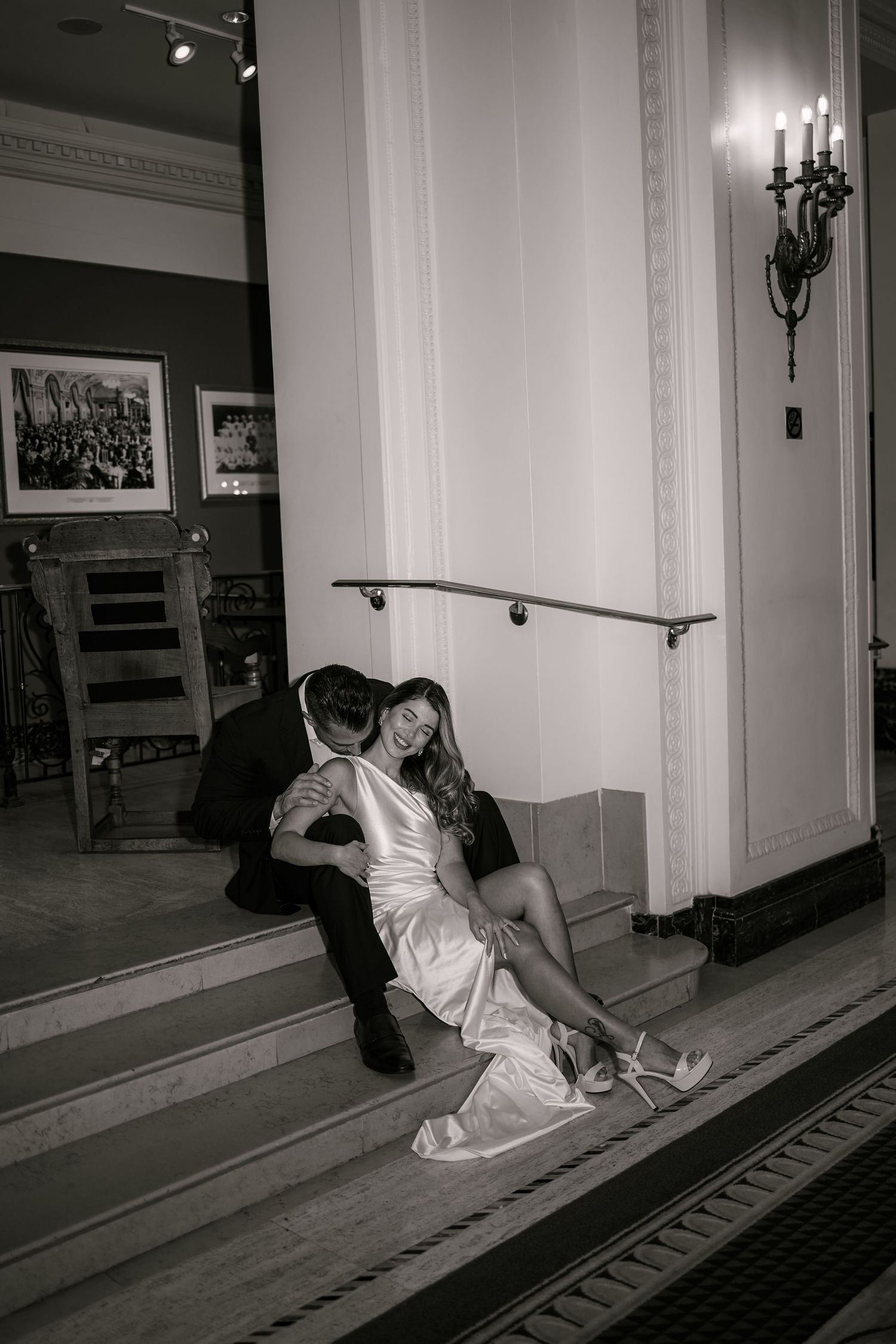 Black and white photo of a bride and groom sitting on elegant staircase steps, sharing a romantic embrace.