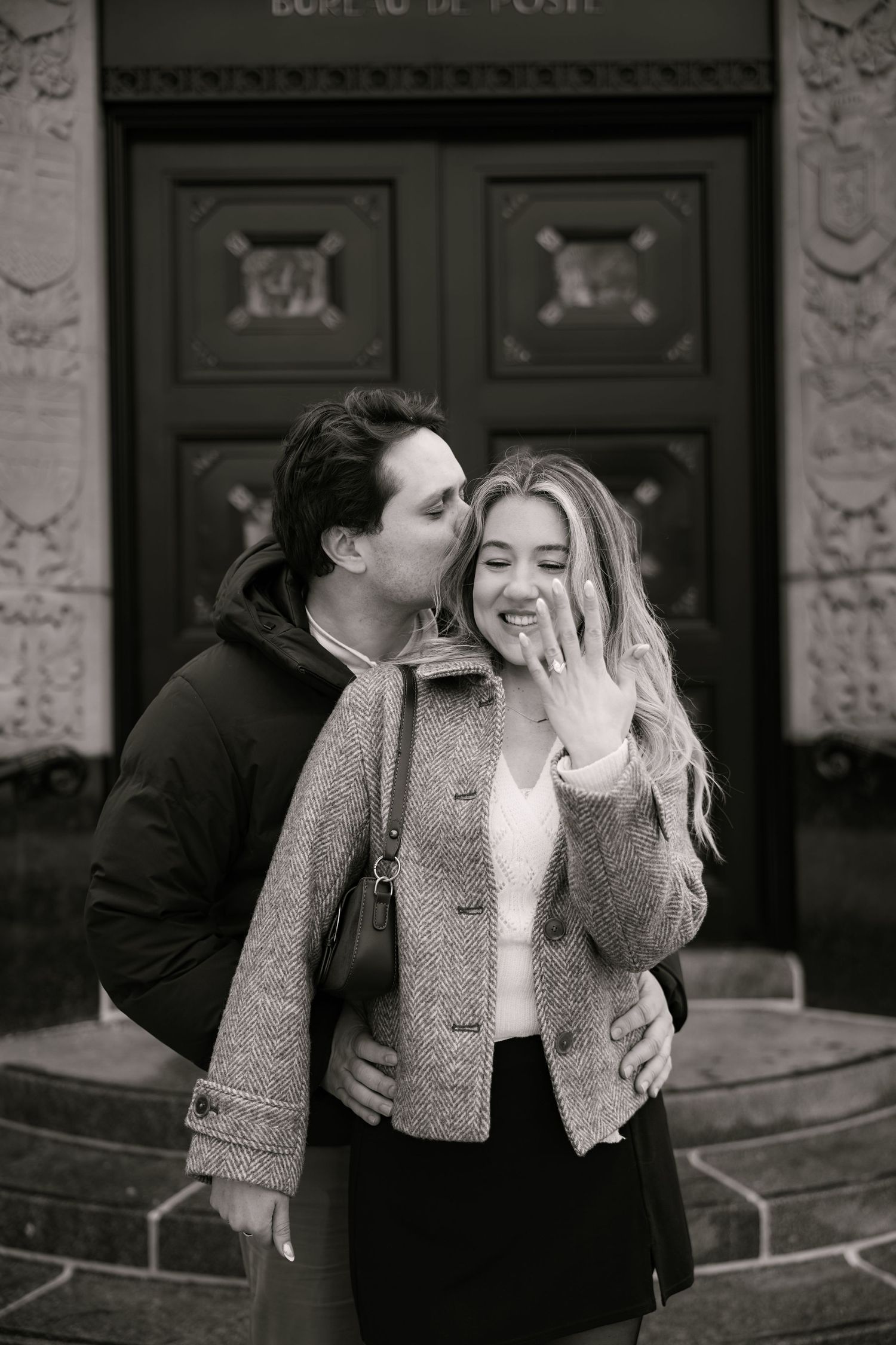 Black and white photo of a couple celebrating engagement, woman showing ring while man kisses her cheek outdoors.