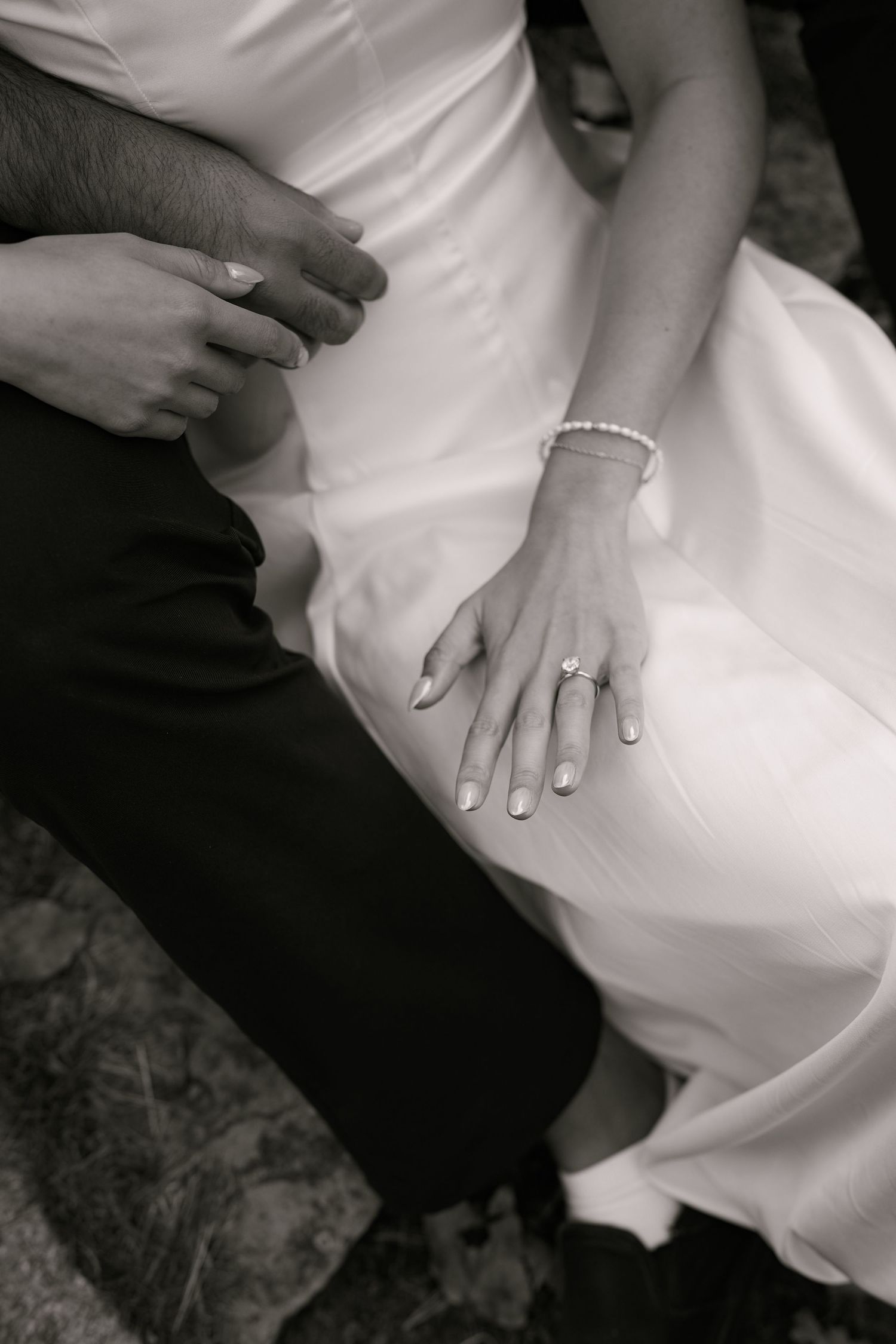 Black and white photo of bride and groom hands showing wedding ring and pearl bracelet on white dress.