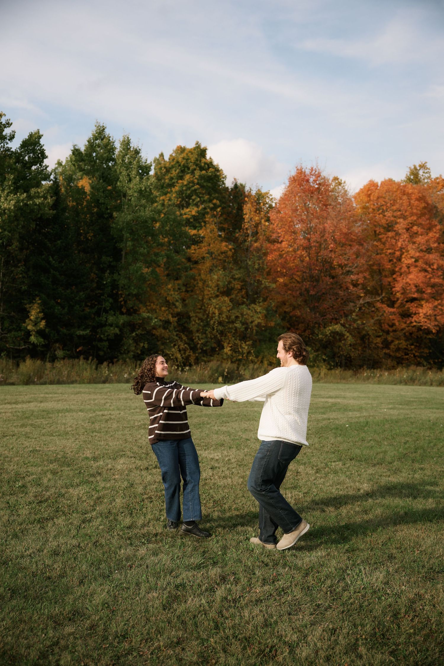 Couple holding hands and dancing in a green field surrounded by colorful autumn trees on a sunny day.