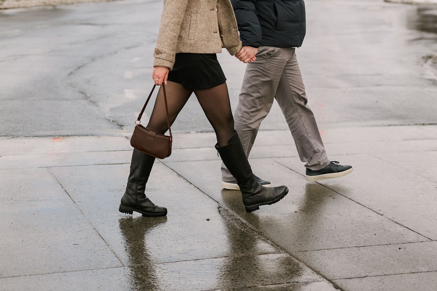 Couple walking on wet sidewalk, woman in black boots holding brown bag, man in gray pants on rainy day.
