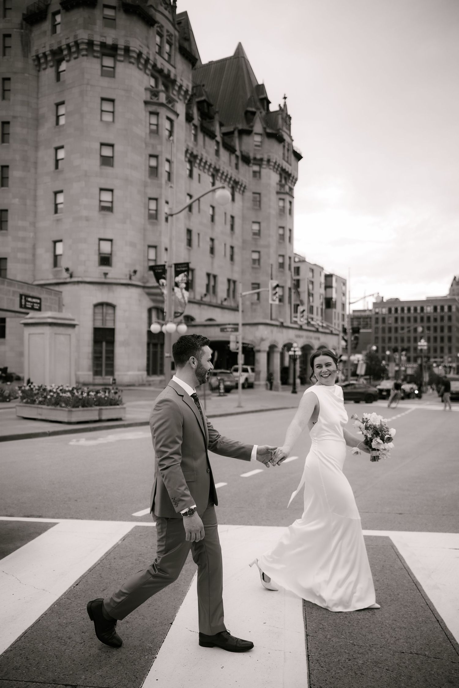 Black and white photo of a bride and groom holding hands while crossing a street in front of a grand historic building.