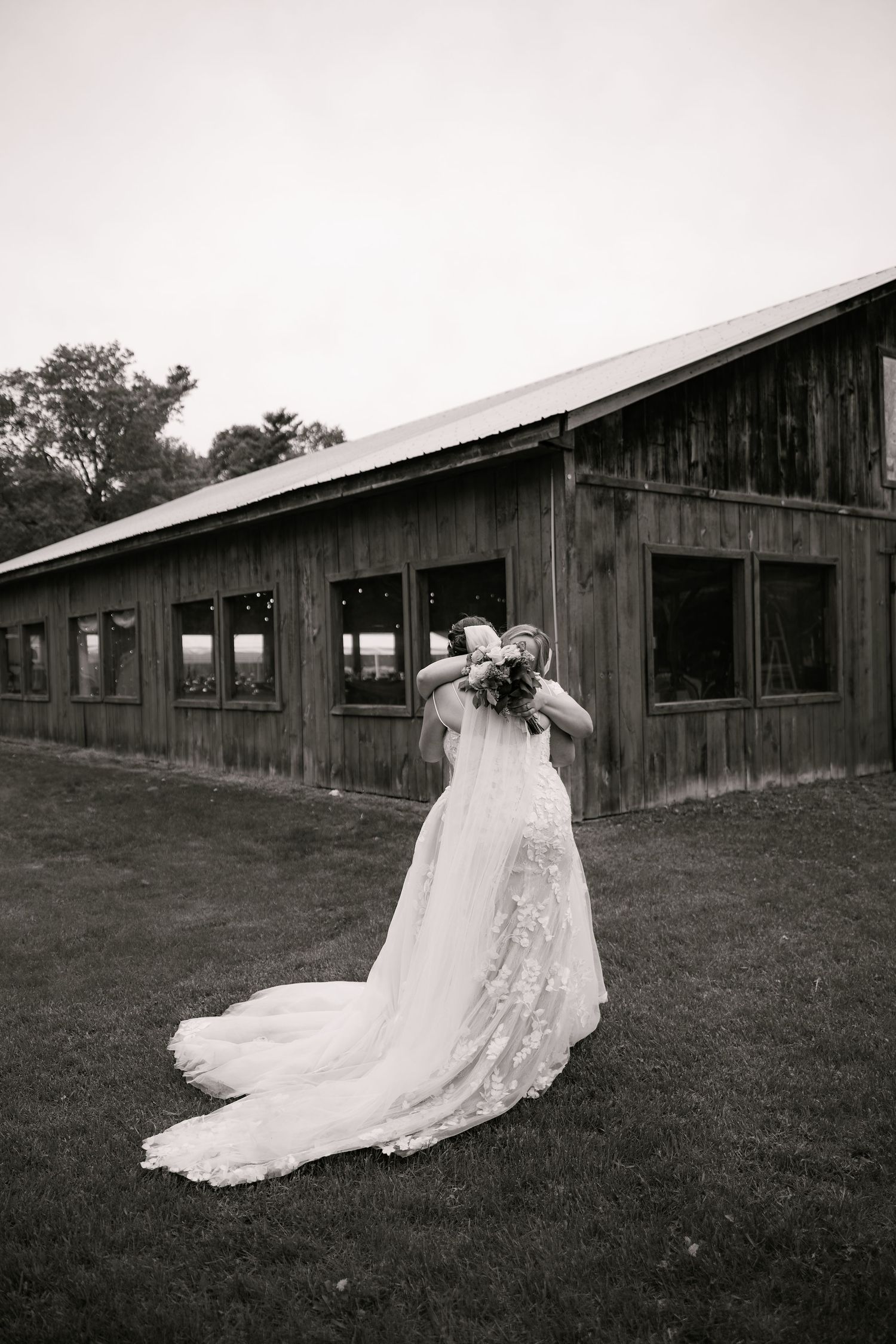 Black and white photo of a bride in a lace gown holding bouquet near a rustic barn on her wedding day.