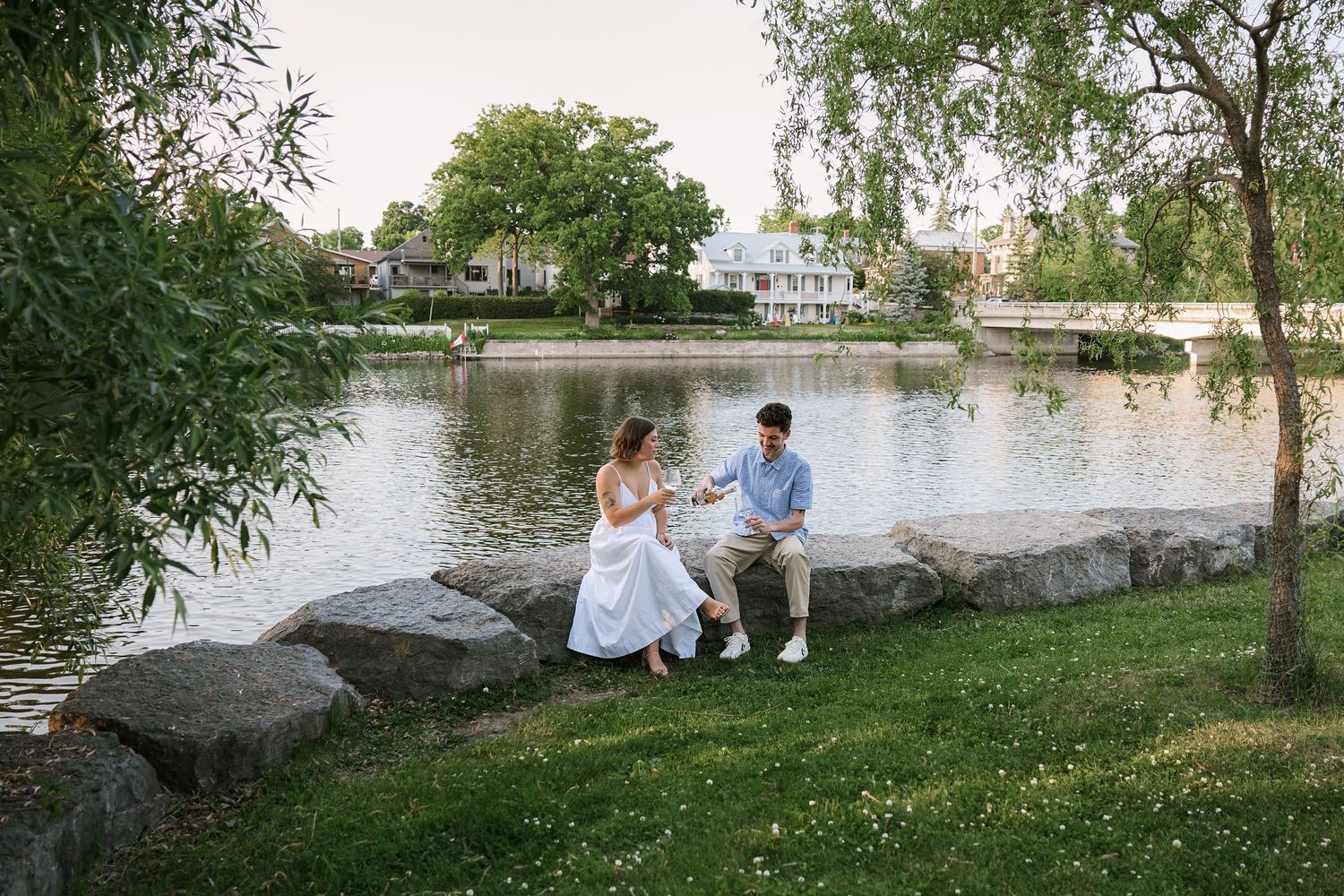 Couple enjoying a romantic picnic by a tranquil lake surrounded by willow trees and rocks on a sunny afternoon.