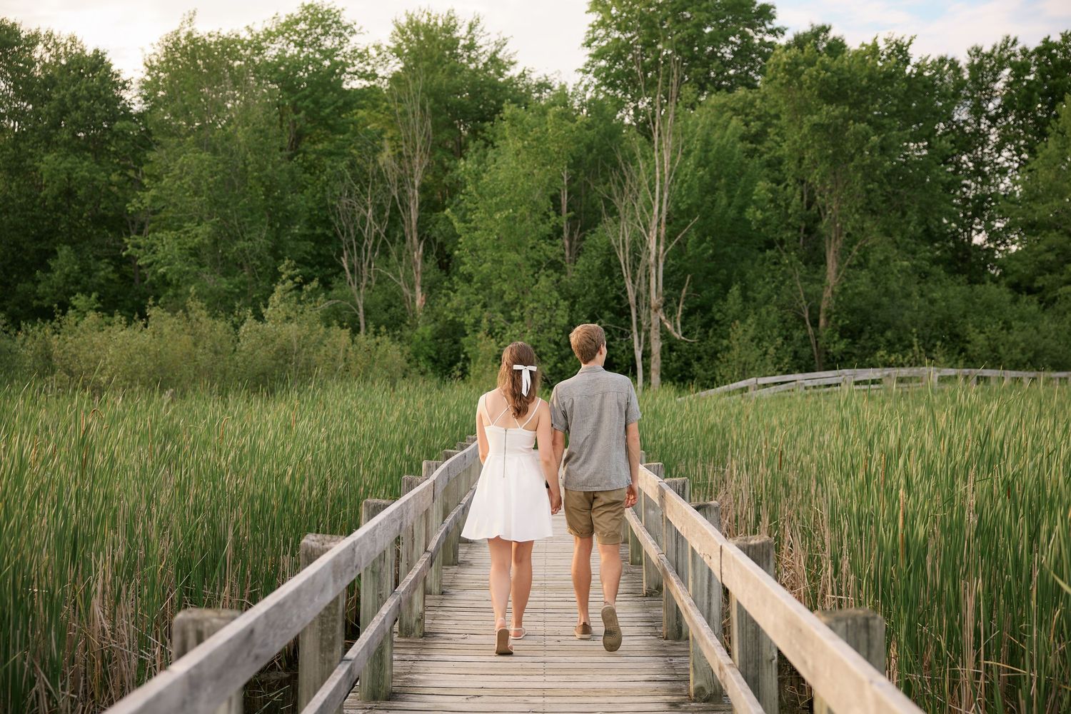 A couple walks hand in hand on a wooden boardwalk through tall marsh grasses.