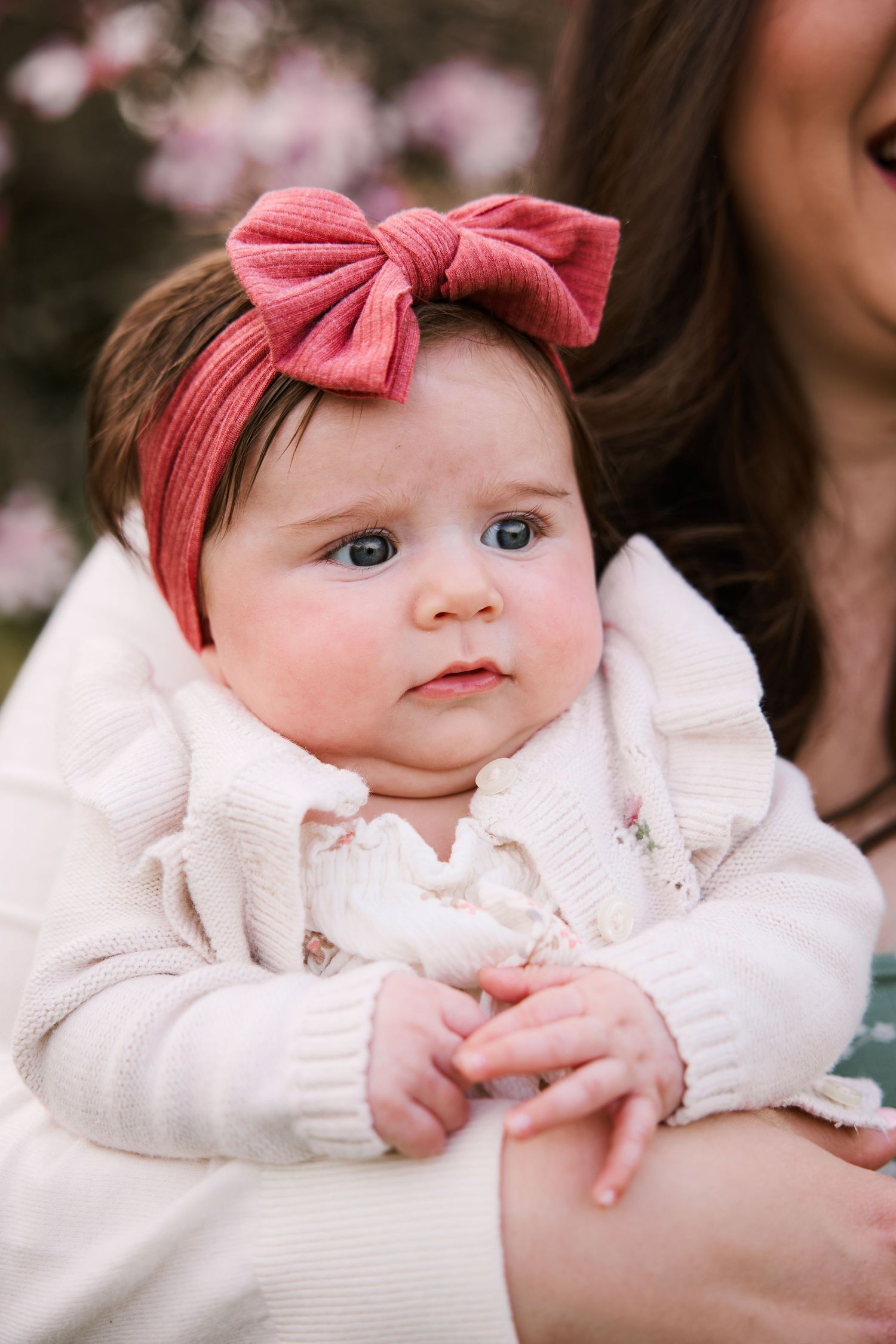 Baby wearing a white sweater and coral bow headband sitting on someone's lap.
