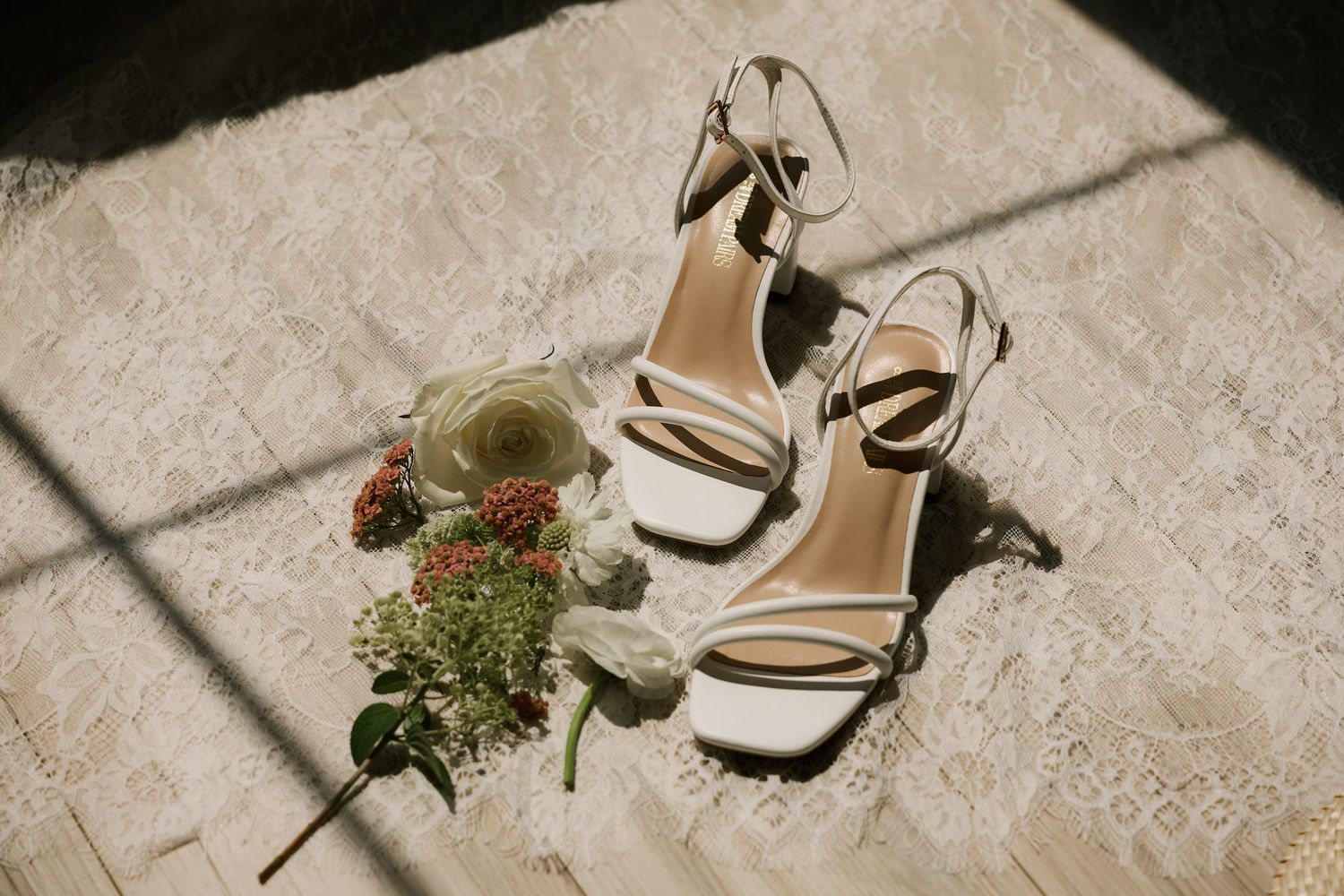 White strappy bridal heels with ankle strap beside fresh flowers and a glass on a lace surface.