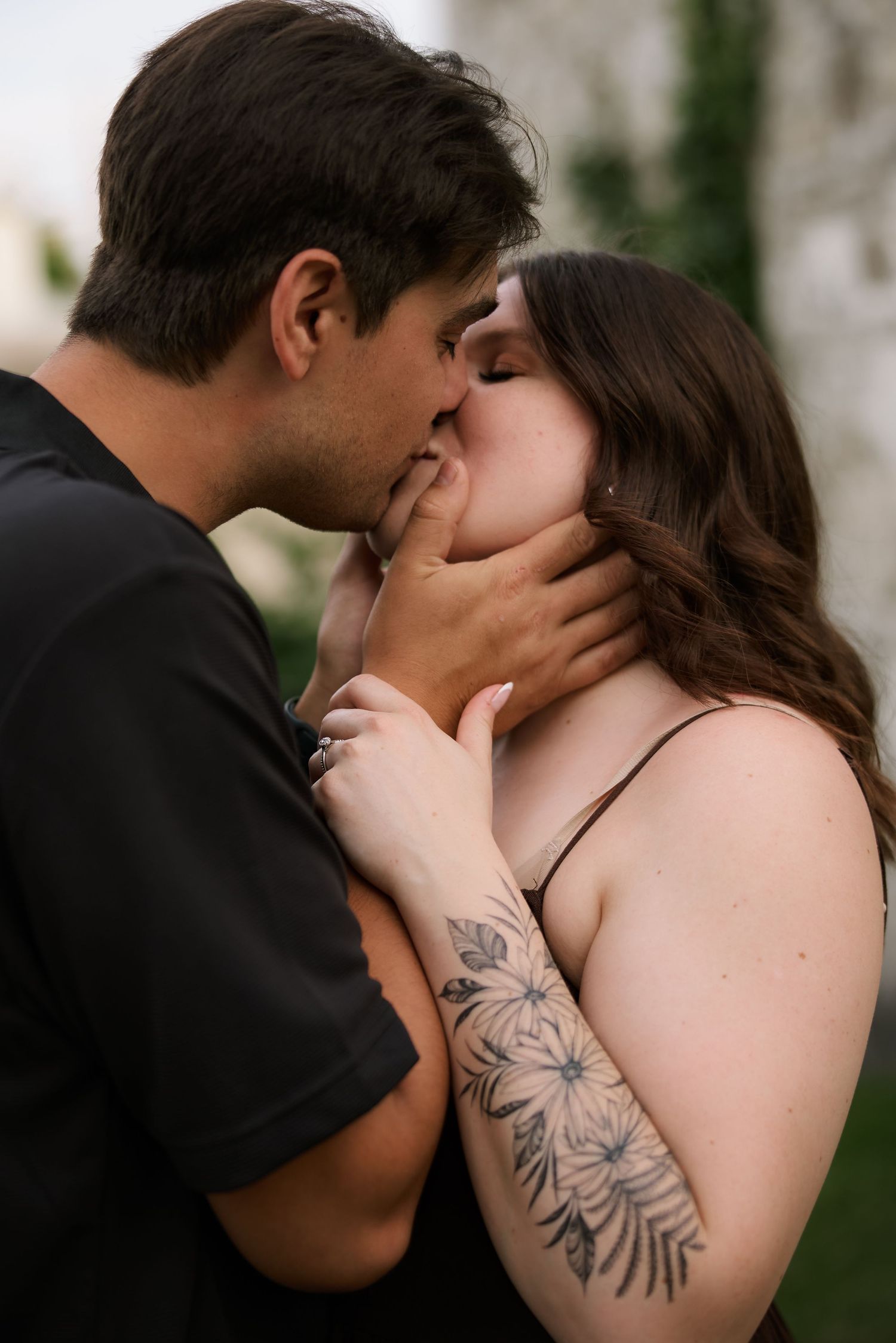 A couple shares a tender kiss outdoors, woman with floral arm tattoo, man gently holding her face.