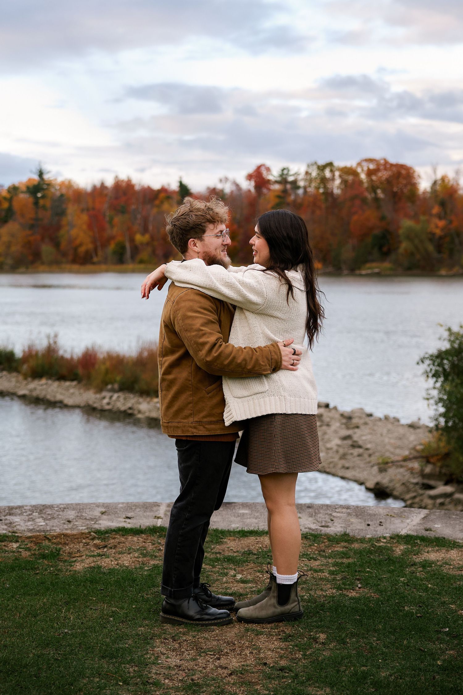 Couple embracing and smiling by a lakeside with colorful autumn foliage trees in the background.