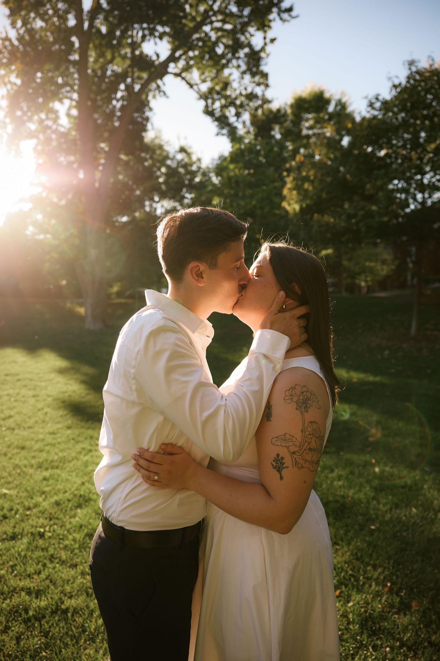 Couple kissing in golden sunlight outdoors, bride in white dress with floral tattoo, groom in white shirt, lush green trees behind.