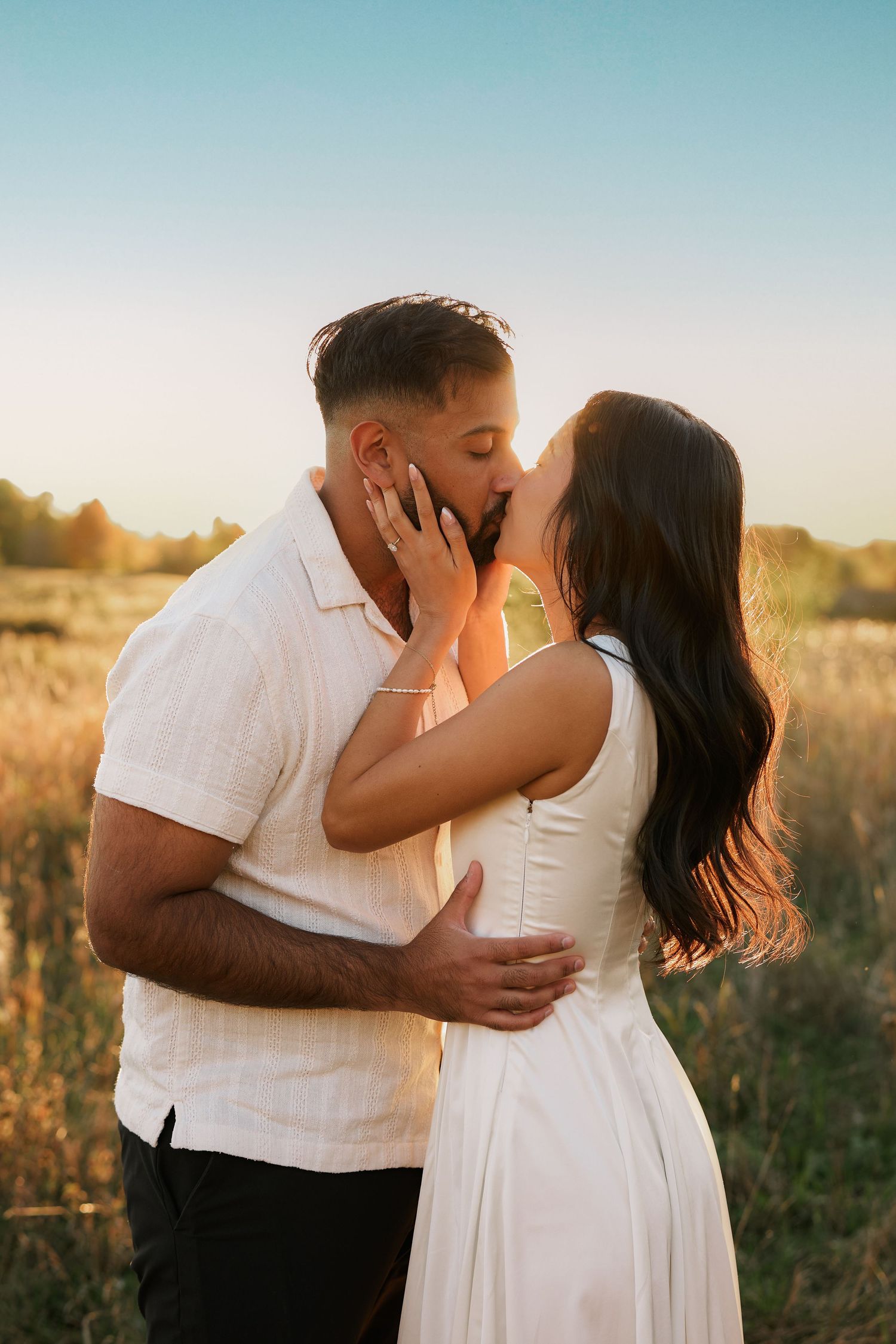 Couple sharing a romantic kiss in a golden field at sunset, woman in white dress, man in white shirt.
