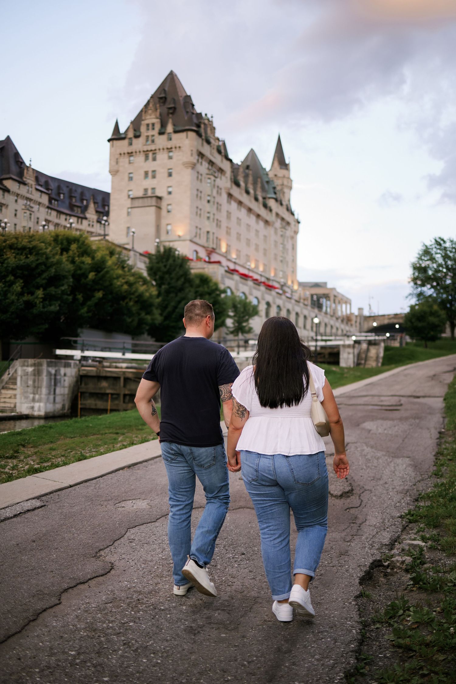 Couple holding hands walking toward a grand château-style hotel at dusk, wearing jeans and casual tops.