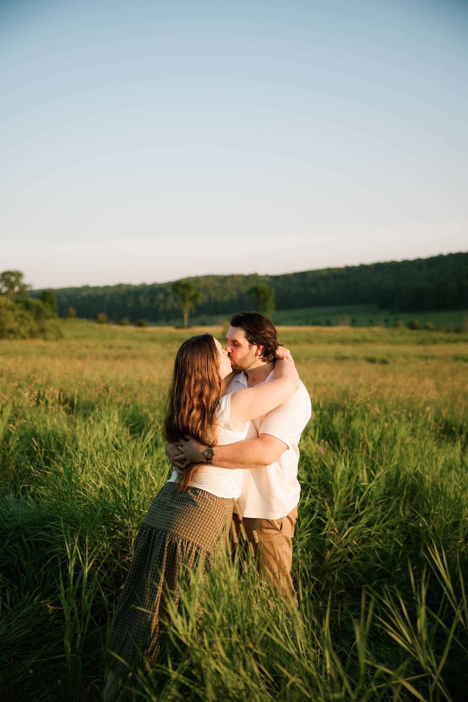 Couple sharing a romantic kiss in a lush green field during golden hour sunset in a scenic countryside.