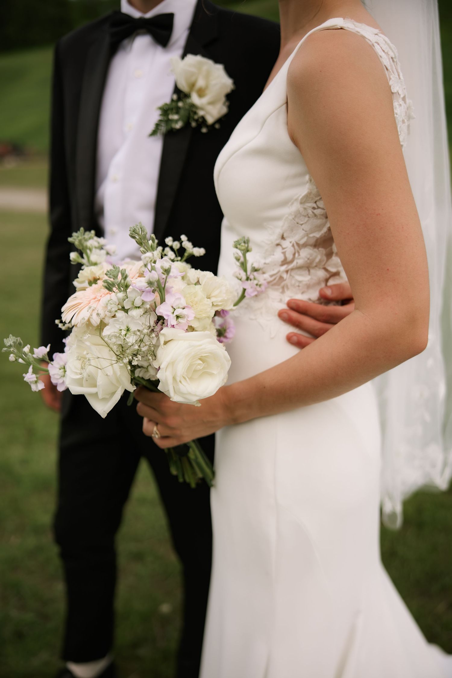 Bride holding white and pink wedding bouquet beside groom in black tuxedo outdoors.
