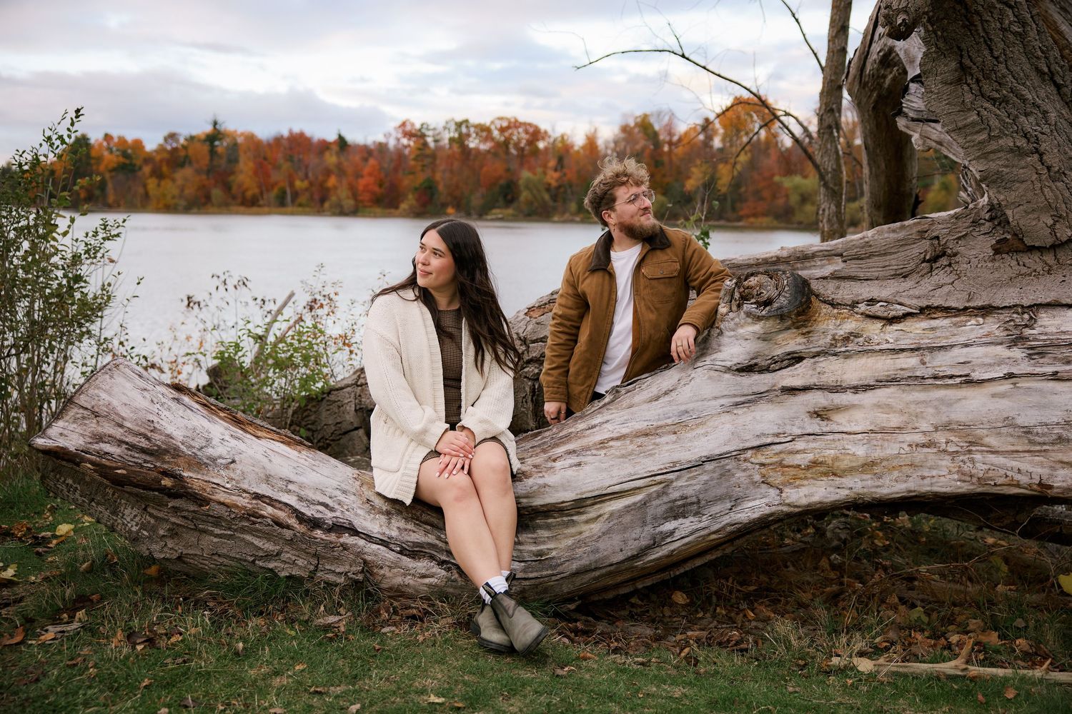 Couple sitting on a fallen log by a lake surrounded by colorful autumn foliage in a romantic outdoor setting.
