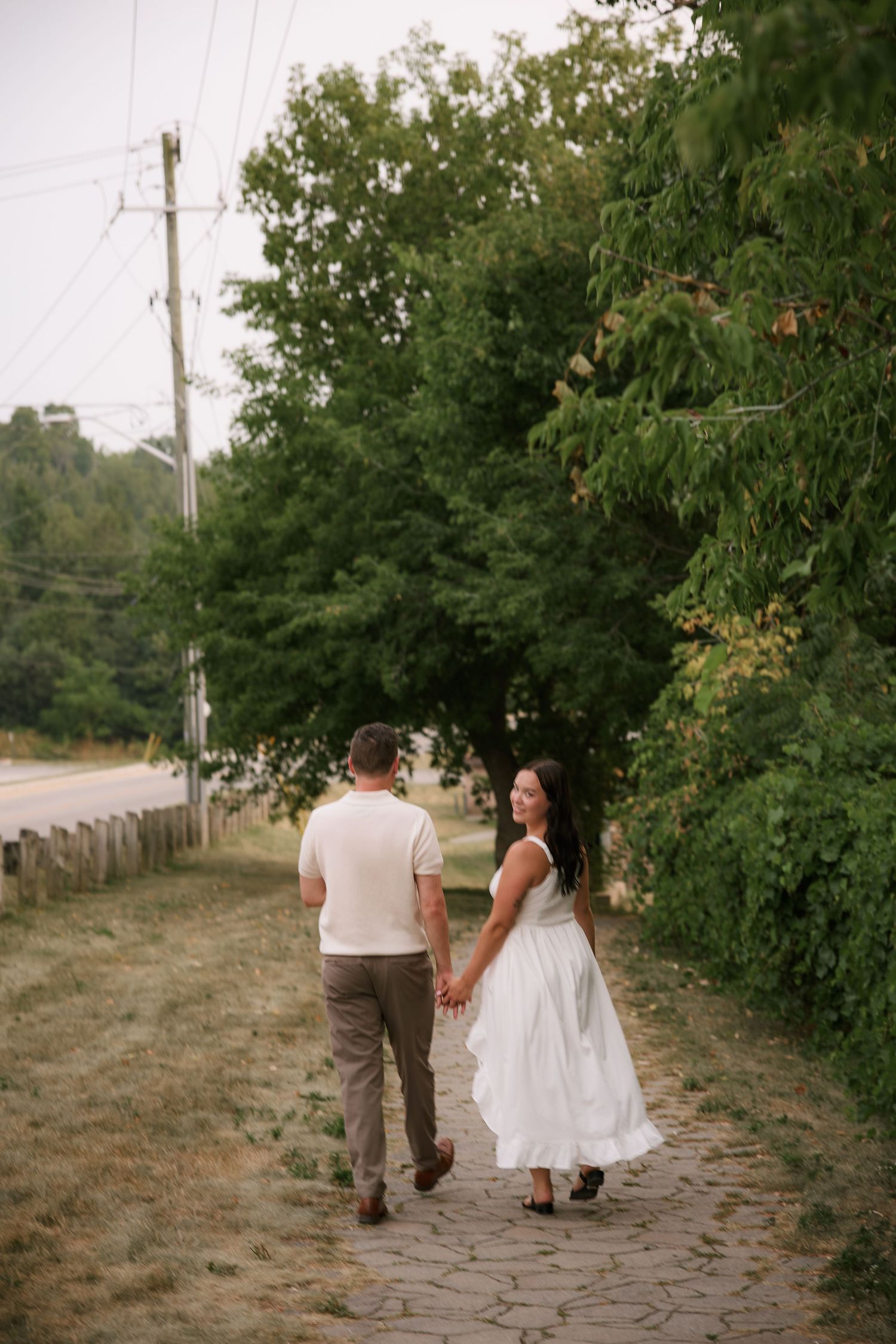 Couple holding hands walking down a stone path surrounded by lush green trees, woman in white dress glancing back.