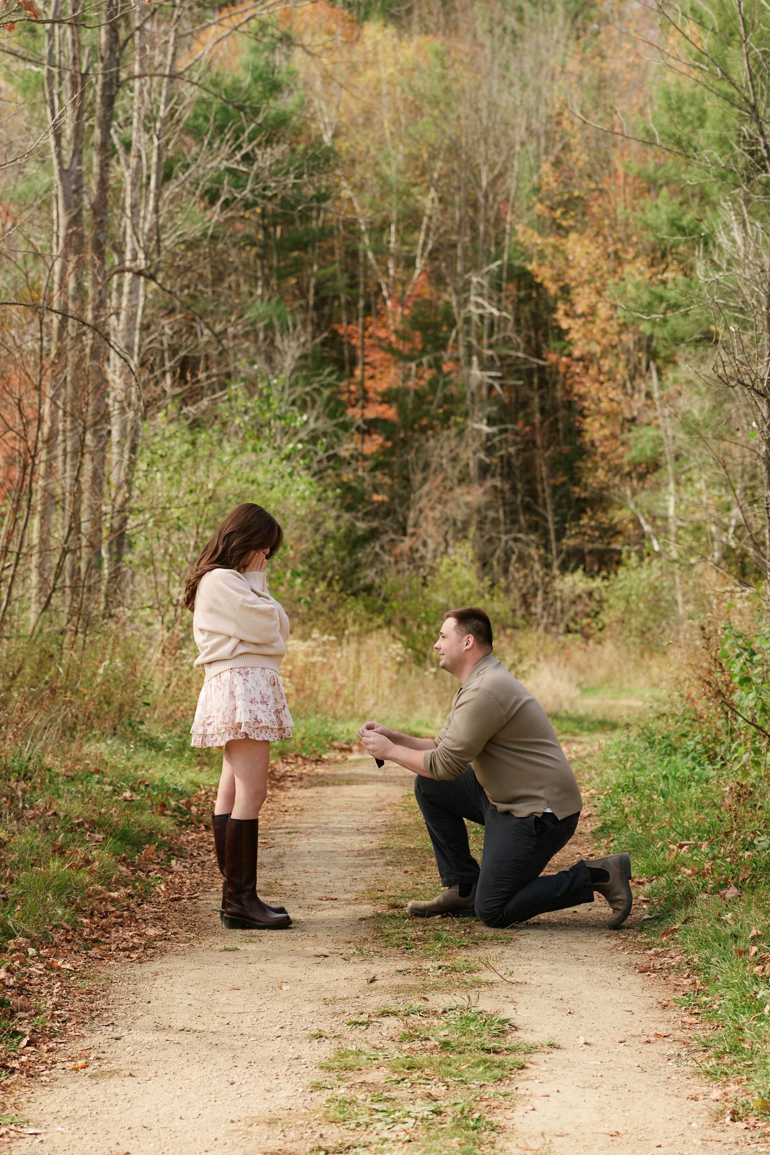 Man proposes on one knee to woman on a scenic autumn forest path surrounded by colorful fall foliage.