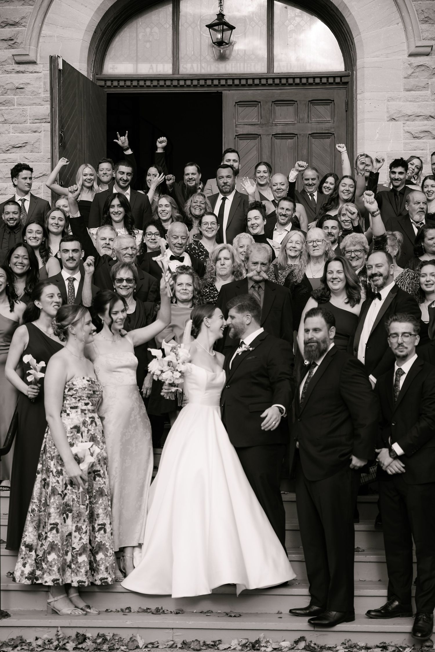 Black and white photo of bride and groom kissing on church steps surrounded by cheering wedding guests.