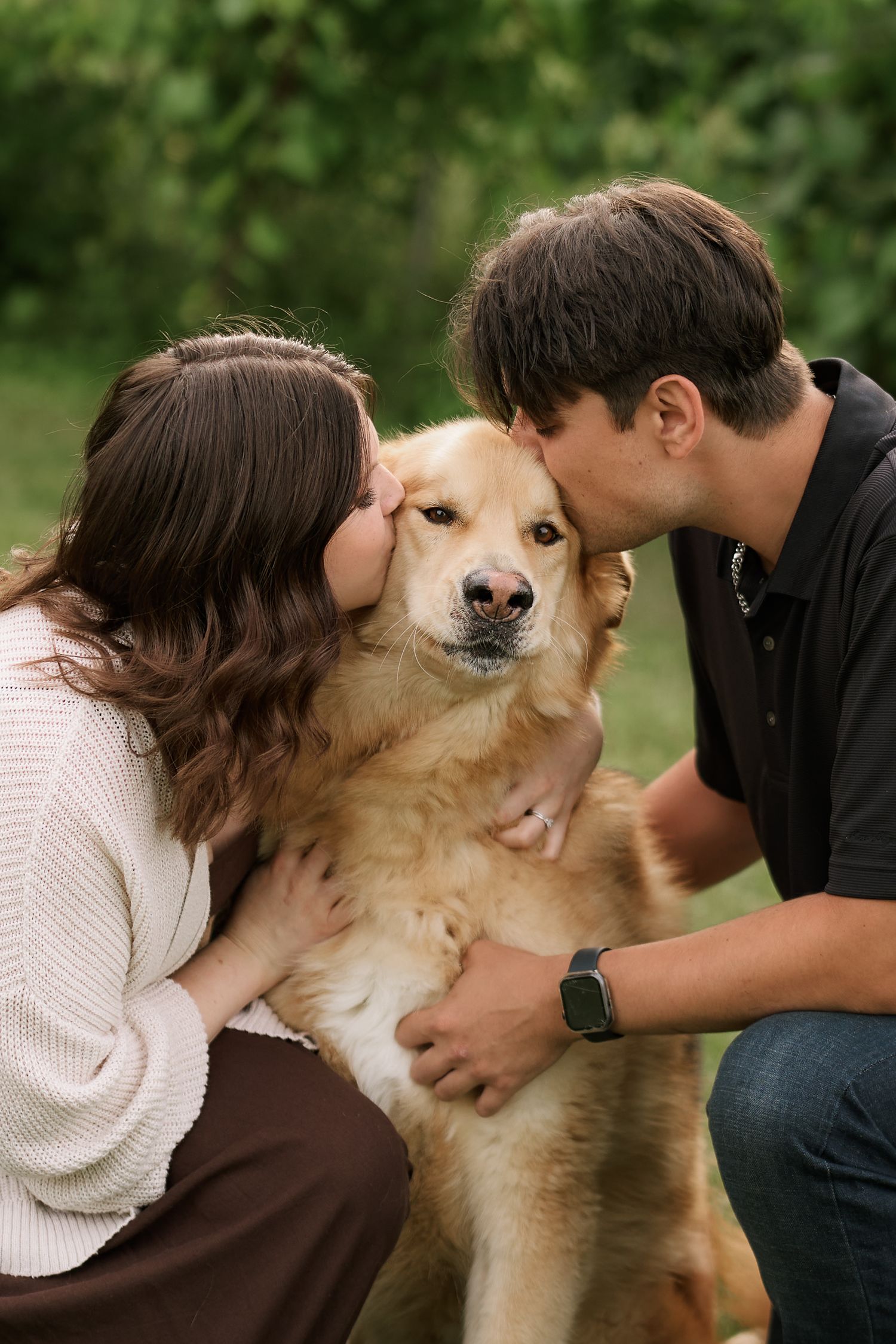 A couple kisses their golden retriever dog on the cheeks outdoors in a green park setting.