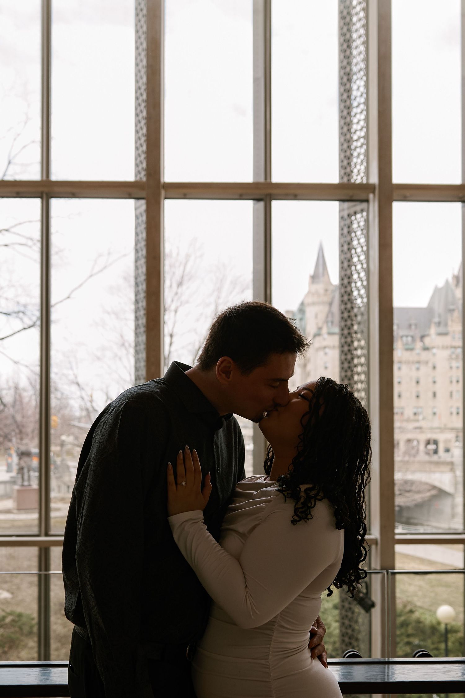 A couple shares a romantic kiss silhouetted against large windows with a historic castle visible outside.