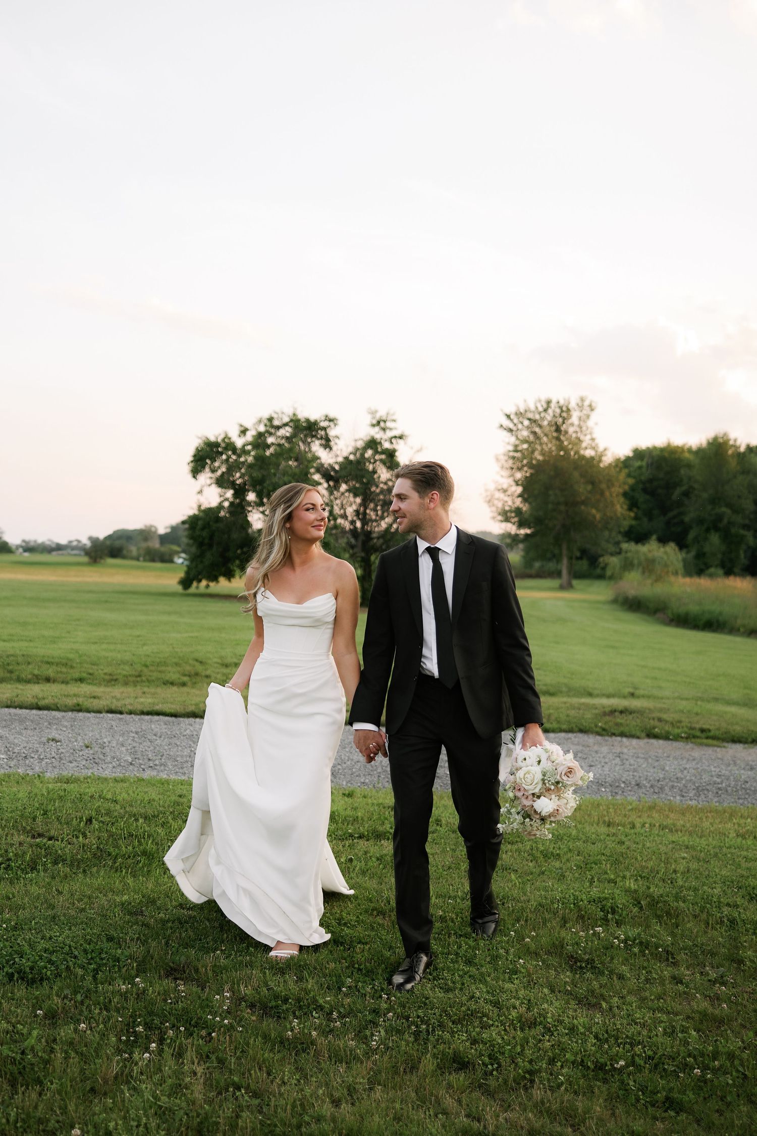 Bride and groom walk hand-in-hand across green field at sunset, she in white gown, he in black suit holding bouquet.