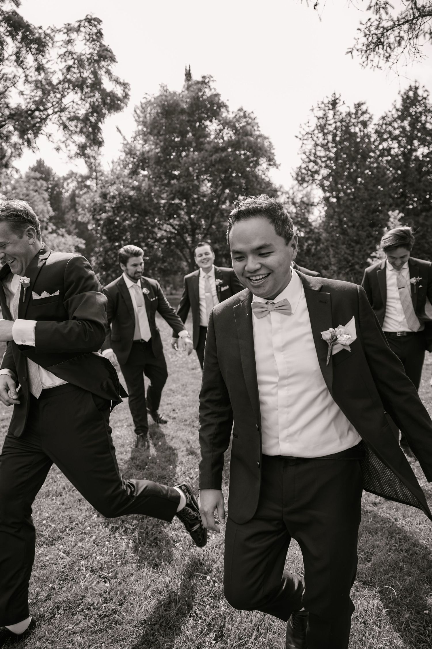 Groomsmen in black suits playfully pose together outdoors in a candid wedding party photo.