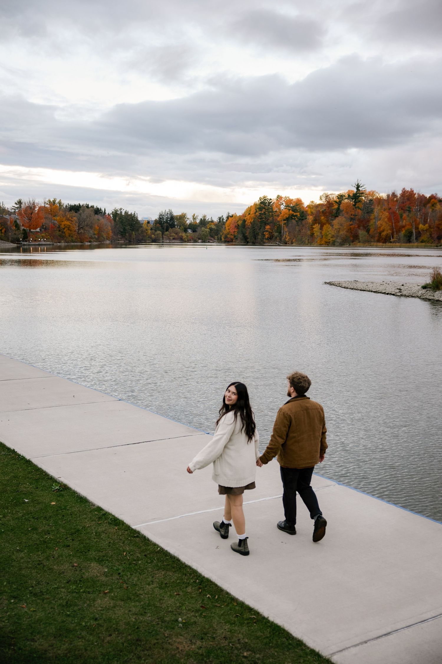 Couple holding hands walking along a lakeside path surrounded by colorful autumn foliage on a cloudy day.