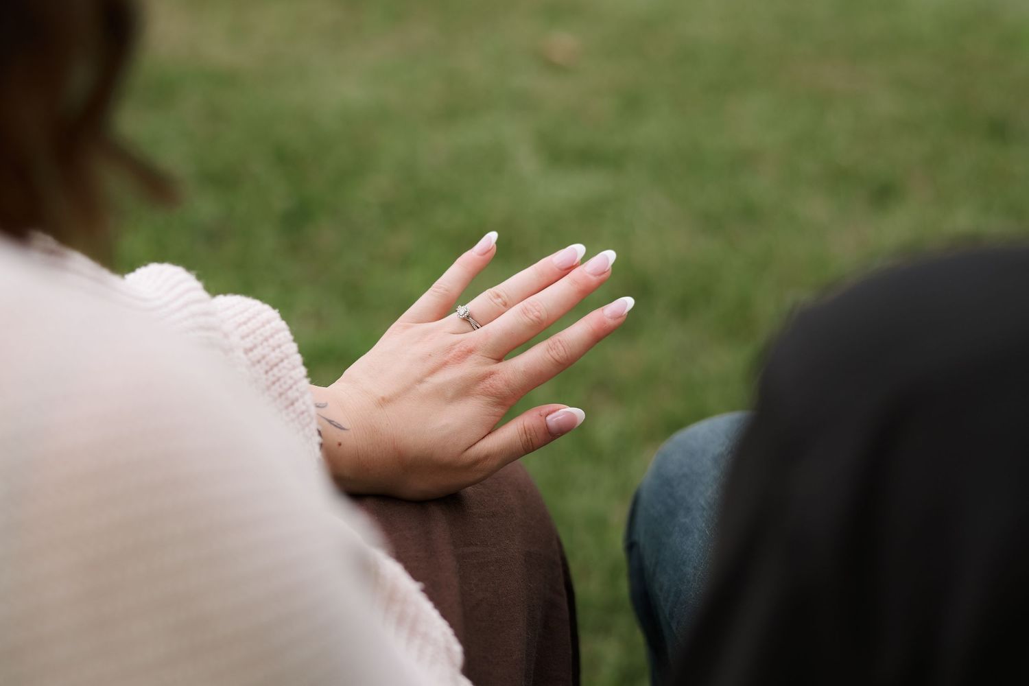 Close-up of a woman's hand displaying a green gemstone ring with light pink manicure, outdoors on grass.