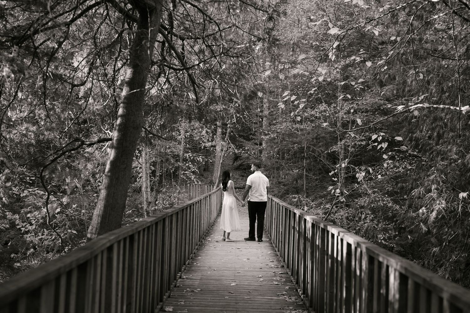 Couple walking hand-in-hand on a wooden bridge surrounded by lush trees in a black and white forest photo.