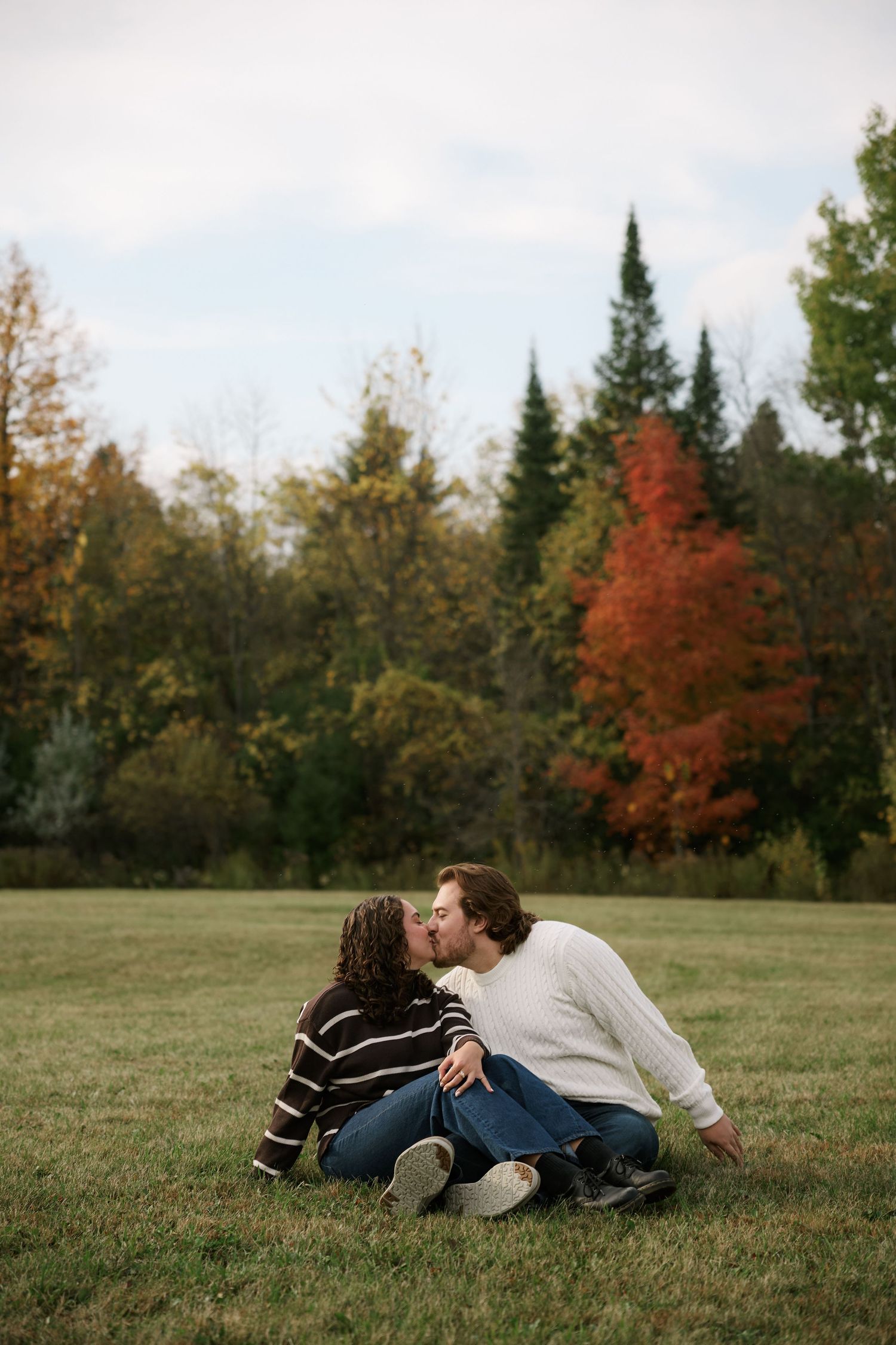 Couple kissing while sitting on grass in a park surrounded by colorful autumn foliage trees.