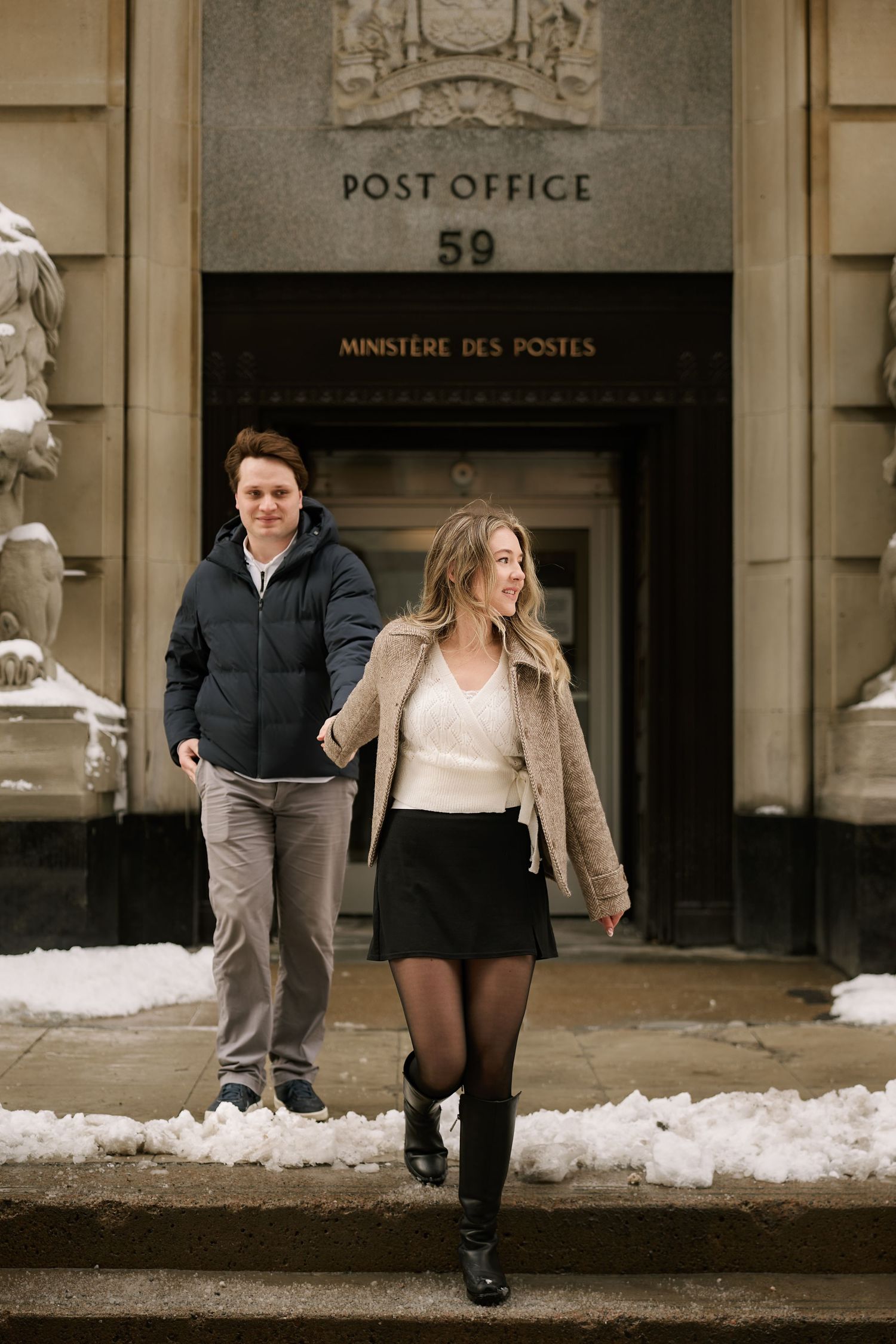 Couple walking out of Post Office 59 in winter, woman in mini skirt and boots, man in puffer jacket, snow on ground.