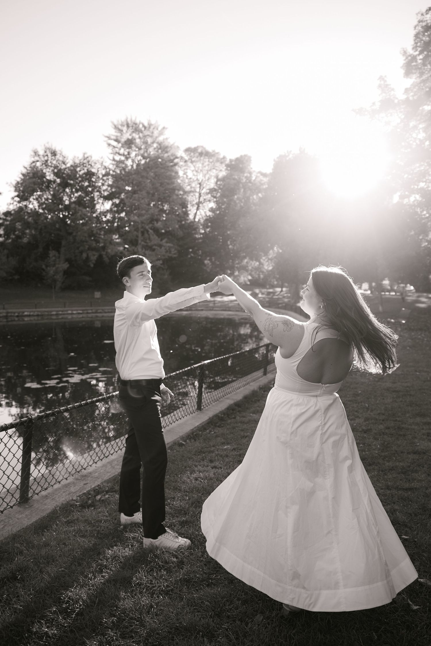 Couple dancing outdoors in black and white photo with sunlight and pond in background.