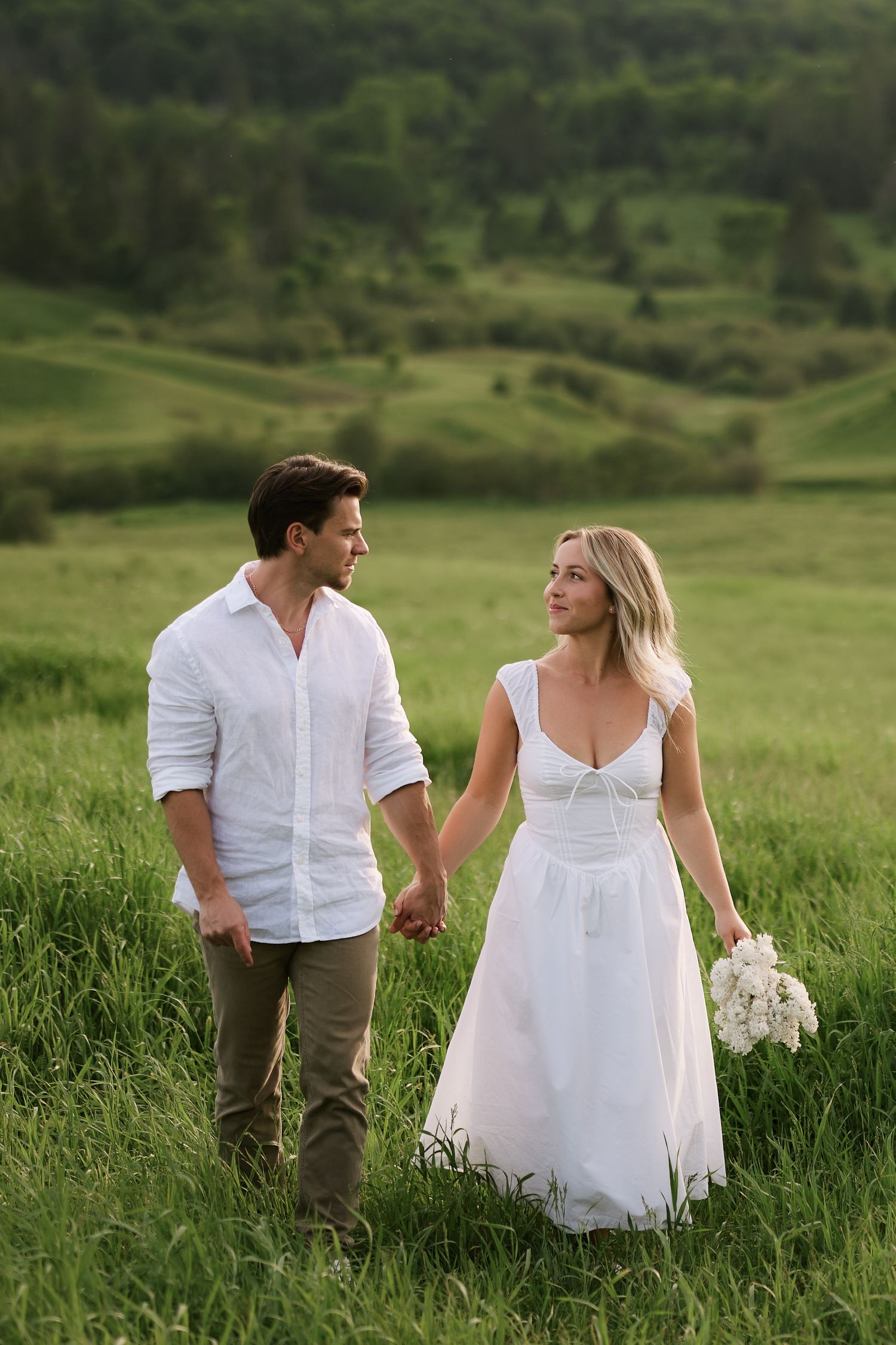 Couple in wedding attire holding hands walking through lush green meadow, bride in white gown holding bouquet.