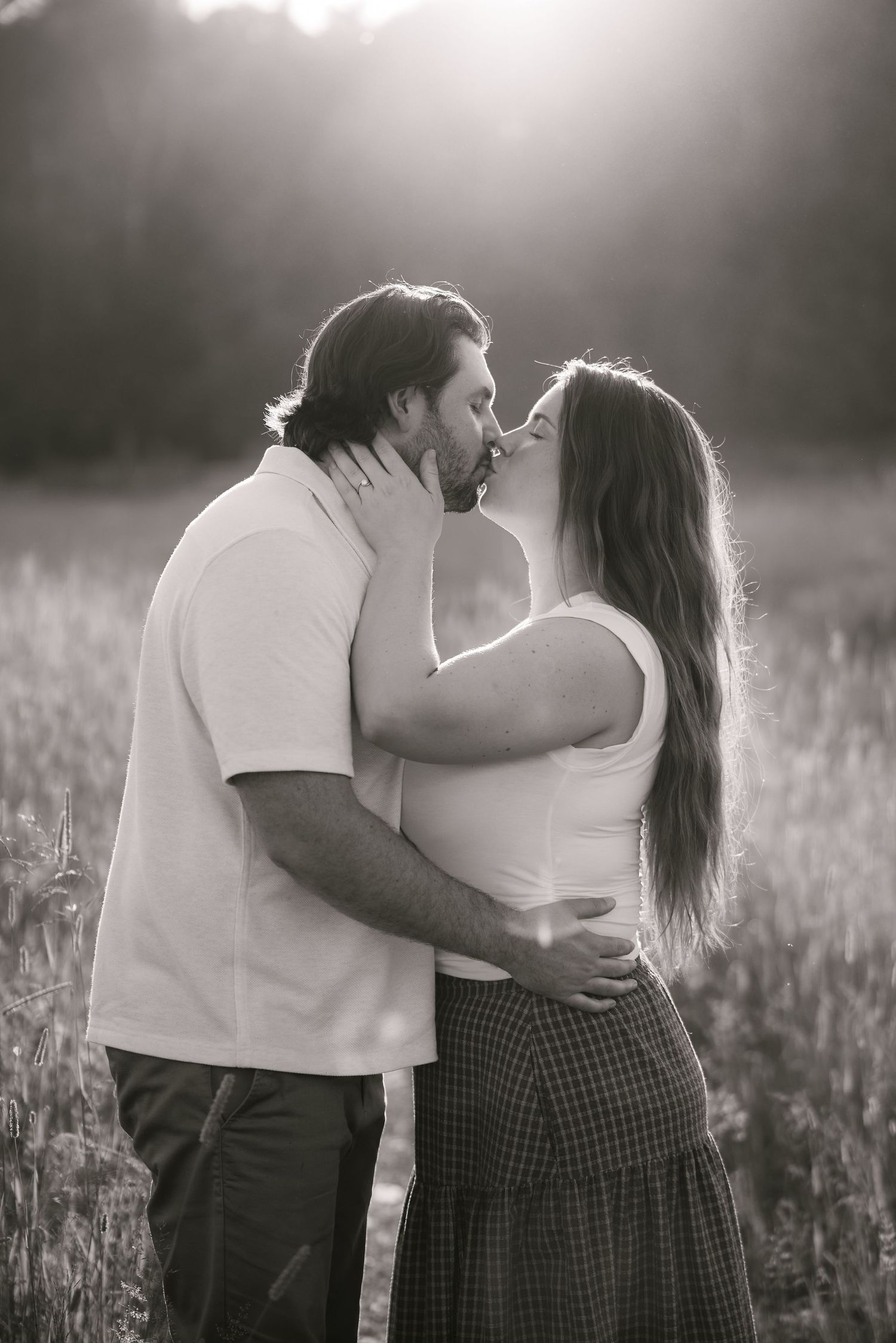 Black and white photo of a couple sharing a tender kiss in a sunlit field, woman with long hair embracing man.