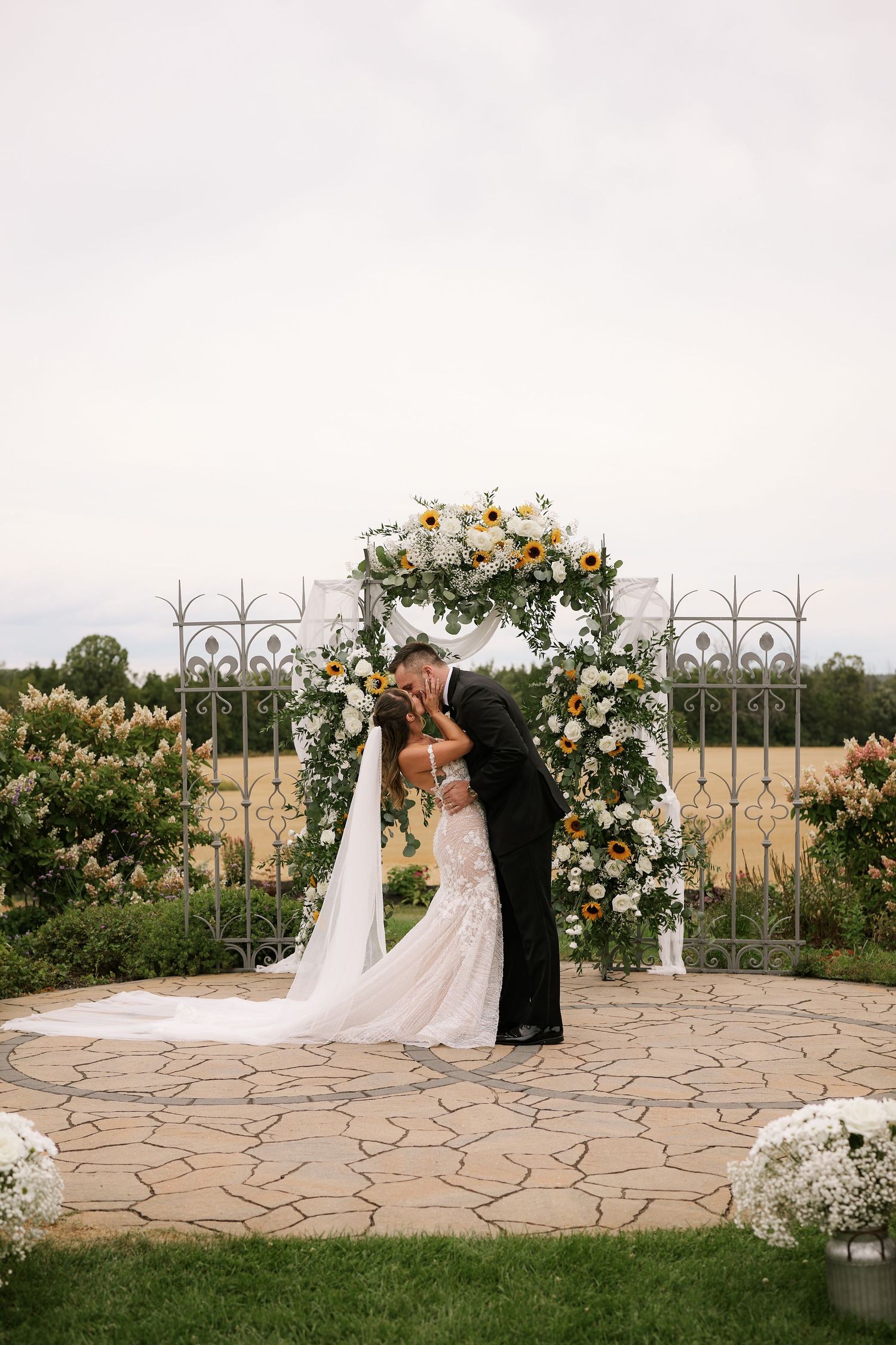 A romantic wedding ceremony takes place on a stone patio with a decorative metal arch and floral arrangements.