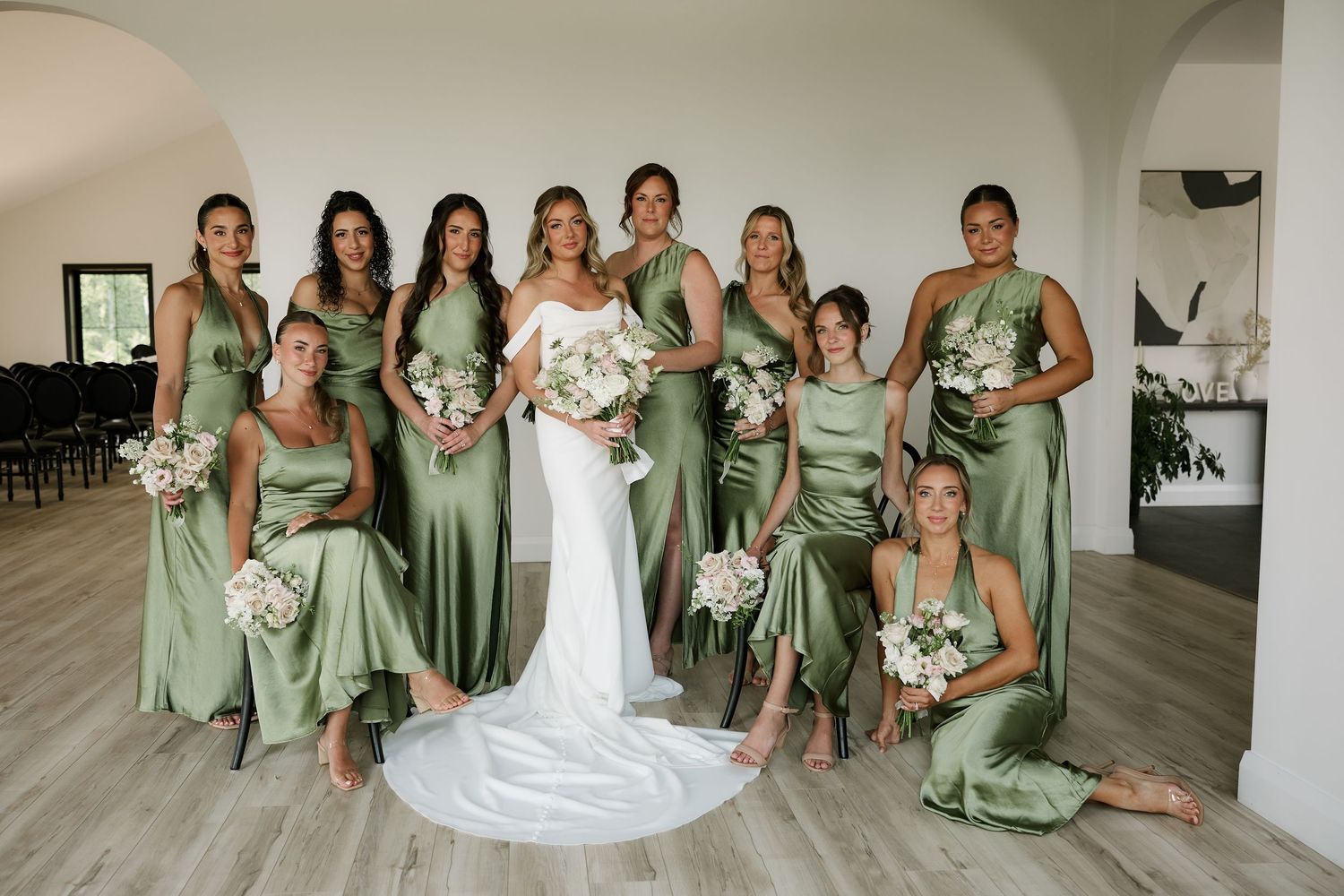 Bride in white gown poses with bridesmaids in sage green dresses holding white bouquets in elegant venue.