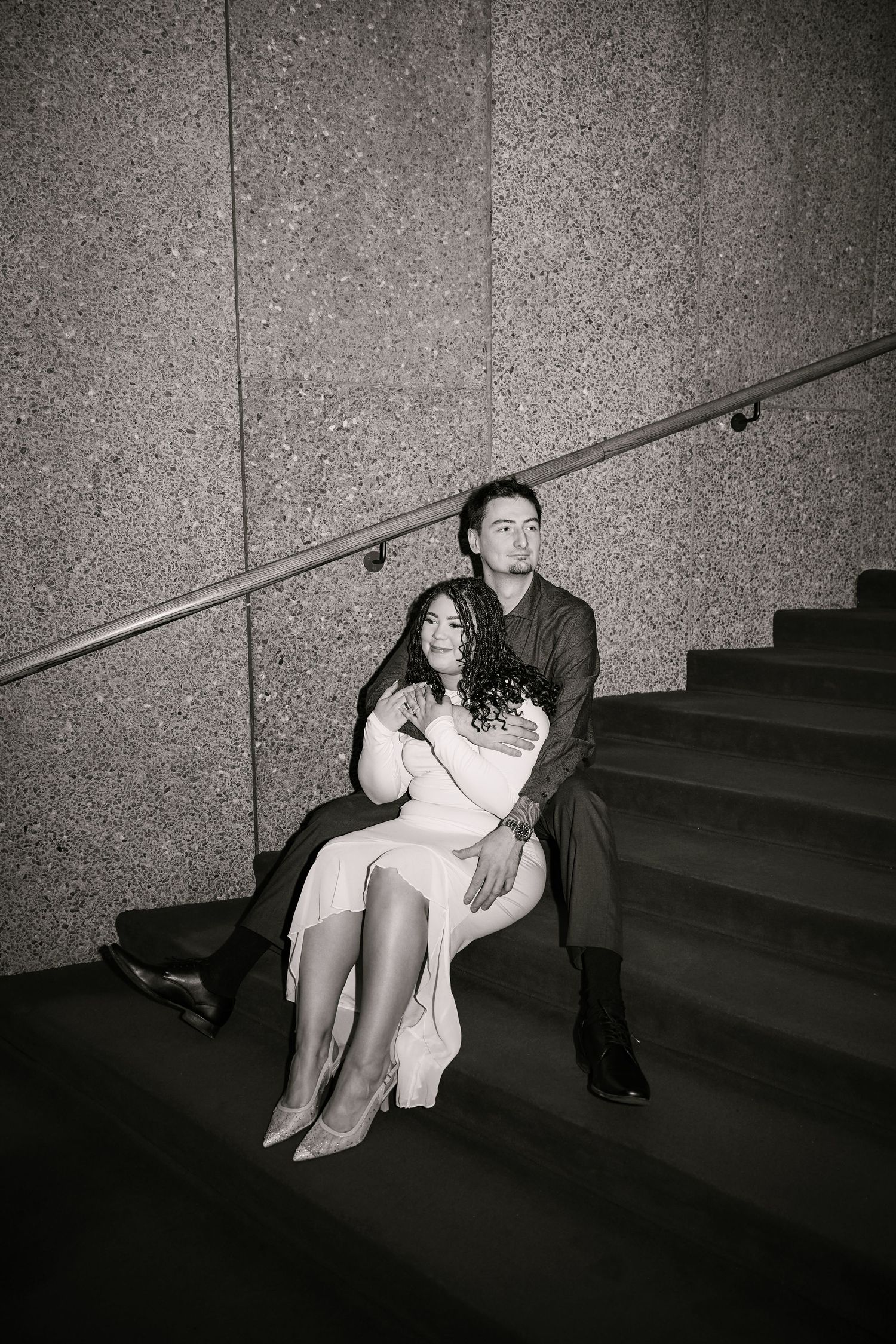Black and white photo of a couple sitting on stairs, woman in dress leaning on man in dark jacket.