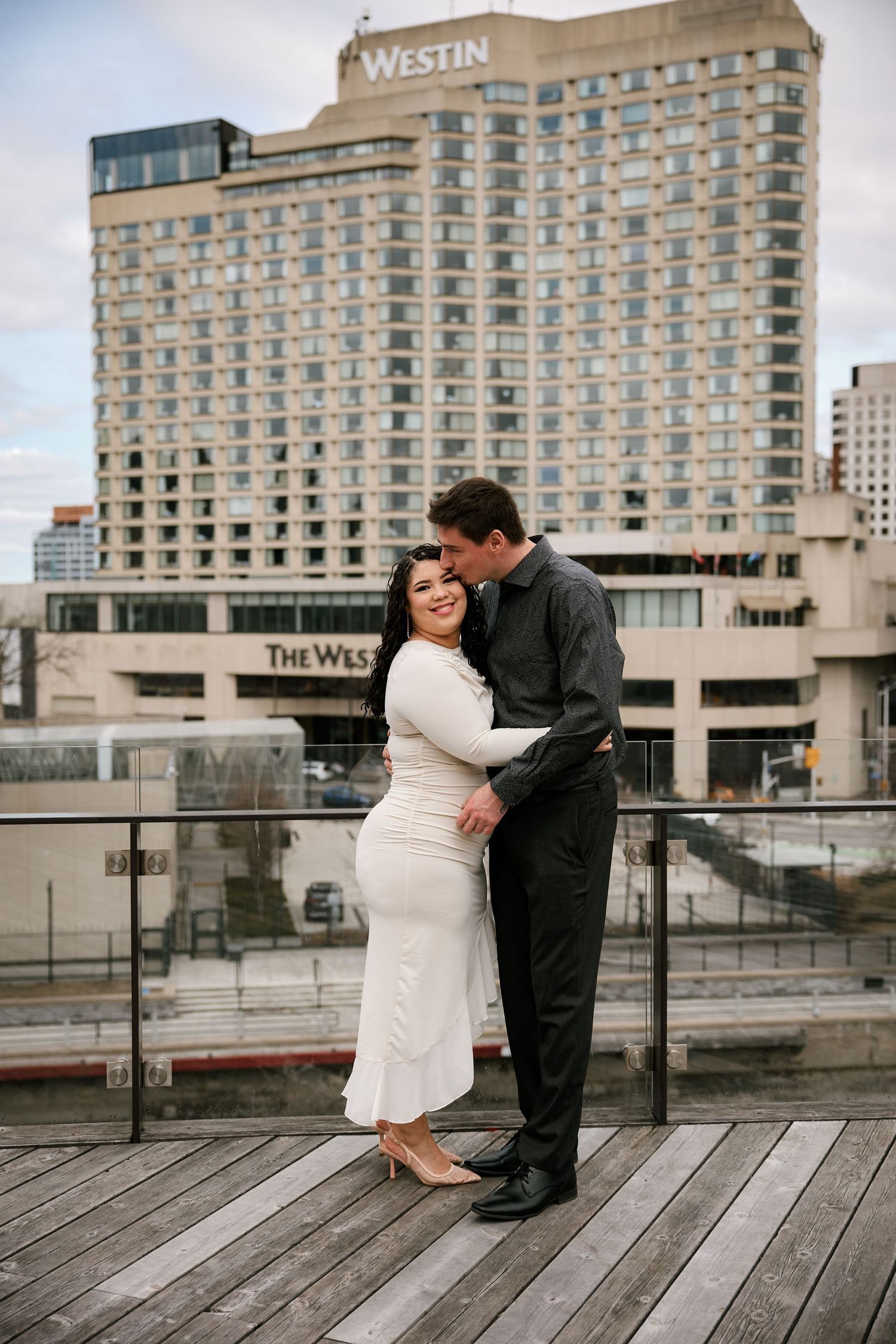 Couple embracing on rooftop deck with The Westin hotel in background, woman in white dress, man in black suit.
