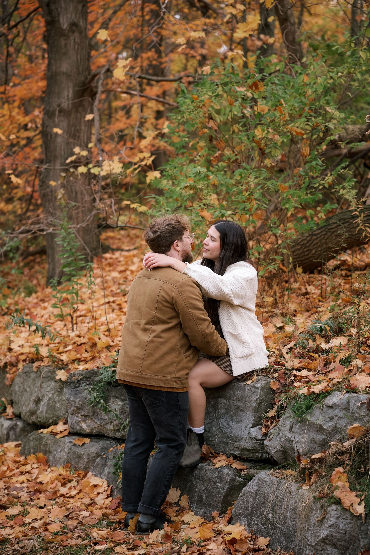 Couple embracing on stone wall in autumn forest, surrounded by vibrant orange and yellow fall foliage.