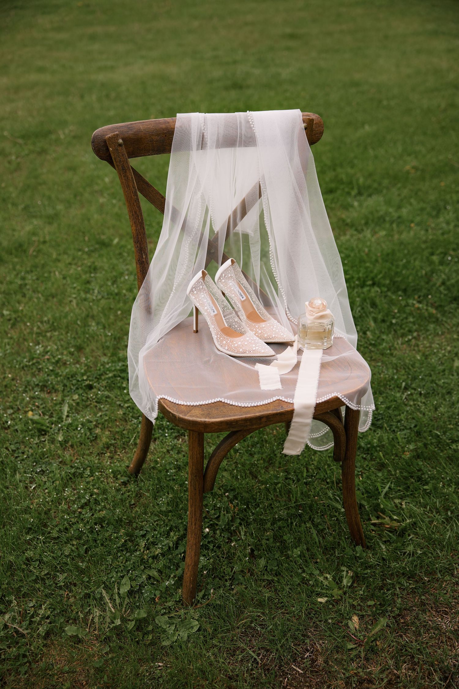 Delicate bridal veil and blush heels arranged on a wooden chair against green grass.