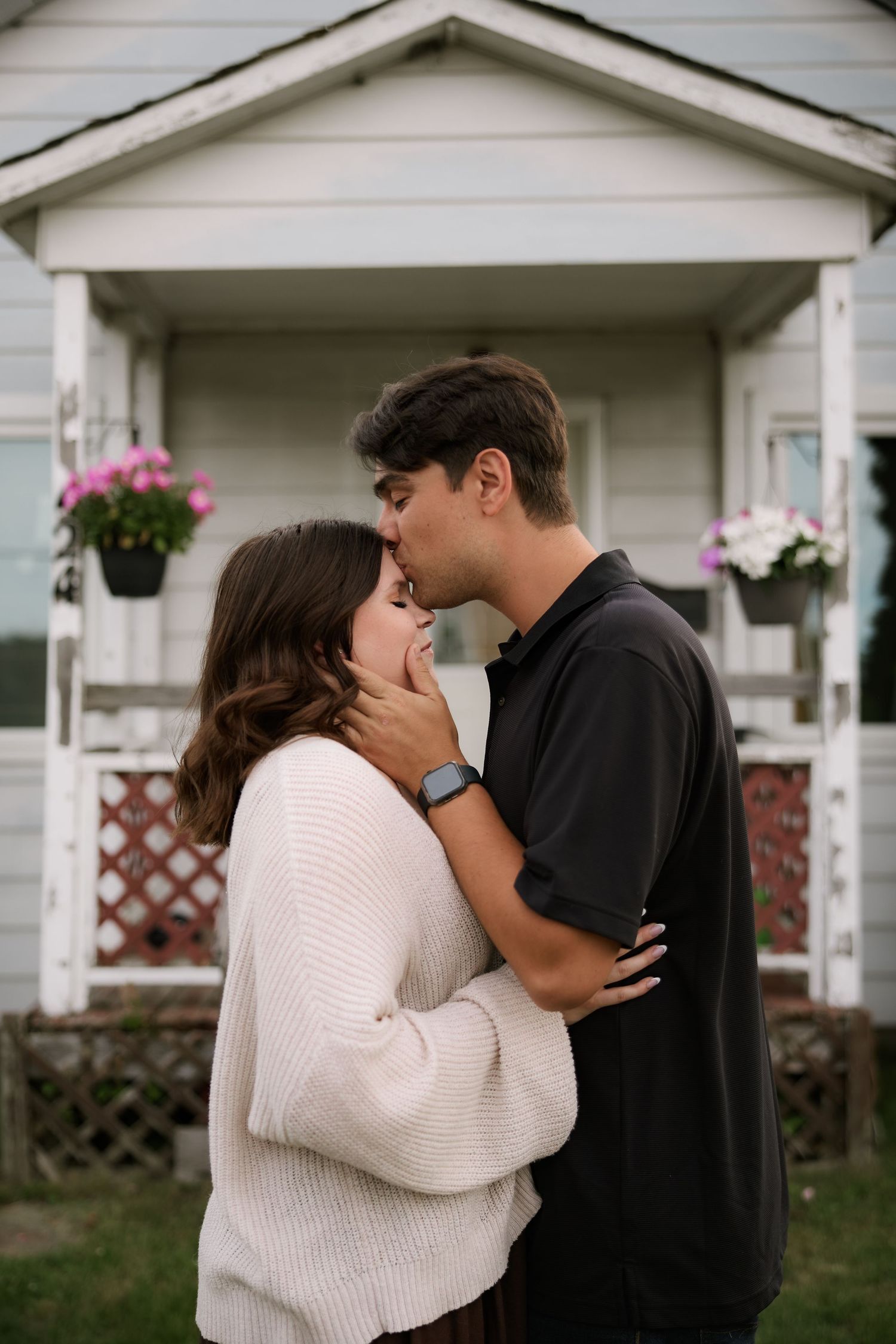 A couple shares a tender kiss in front of a charming white house with flower-adorned porch.