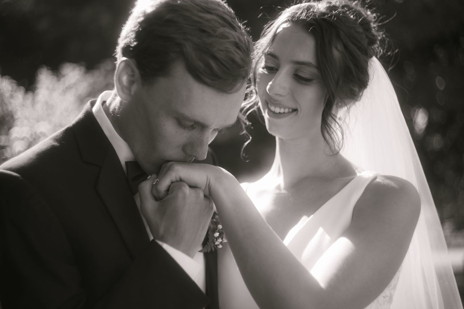 Black and white wedding photo of groom kissing bride's hands while she smiles tenderly, wearing veil and bridal gown.