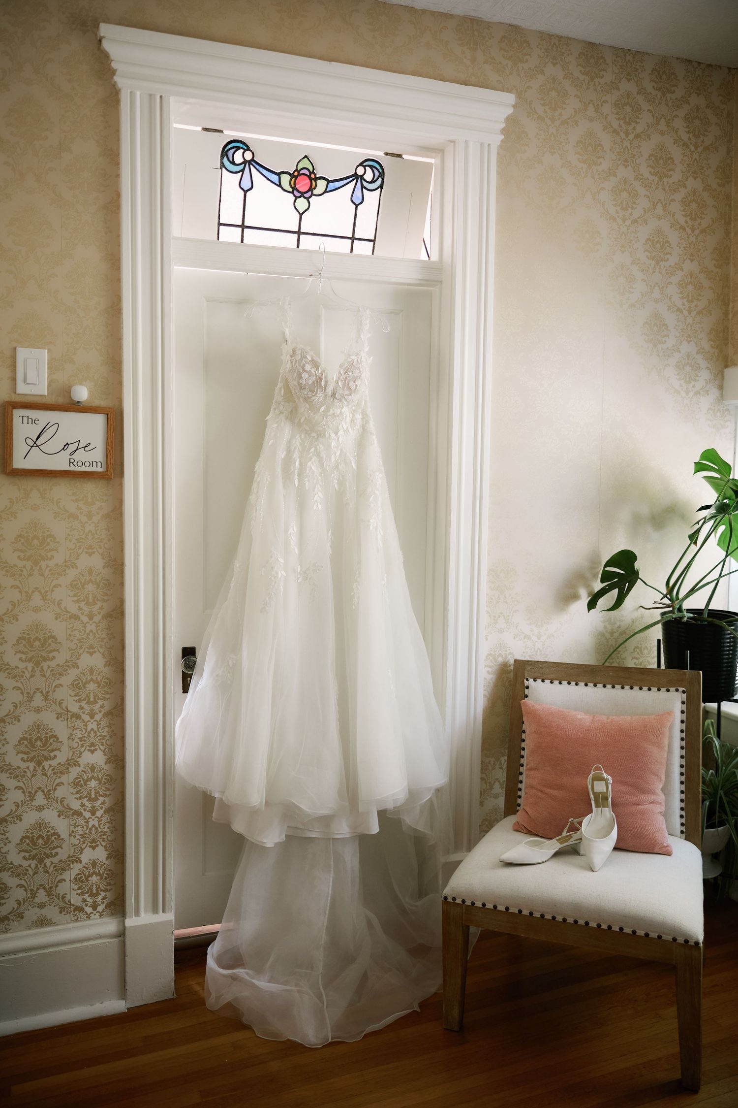 White lace wedding dress hanging in doorway with stained glass window, bridal heels on chair with pink pillow in elegant room.
