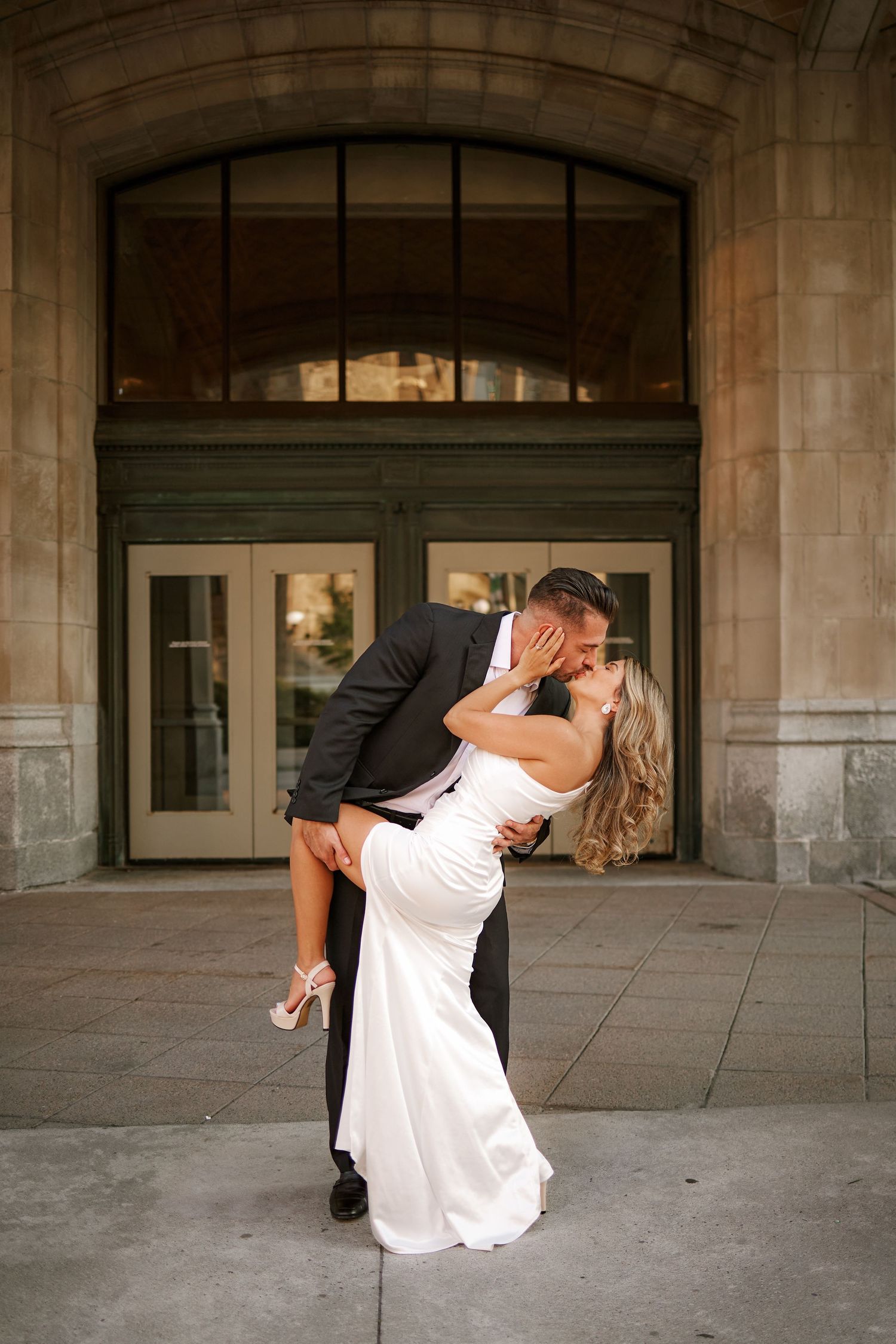 Bride and groom share a romantic dip kiss in front of elegant stone building doors on their wedding day.