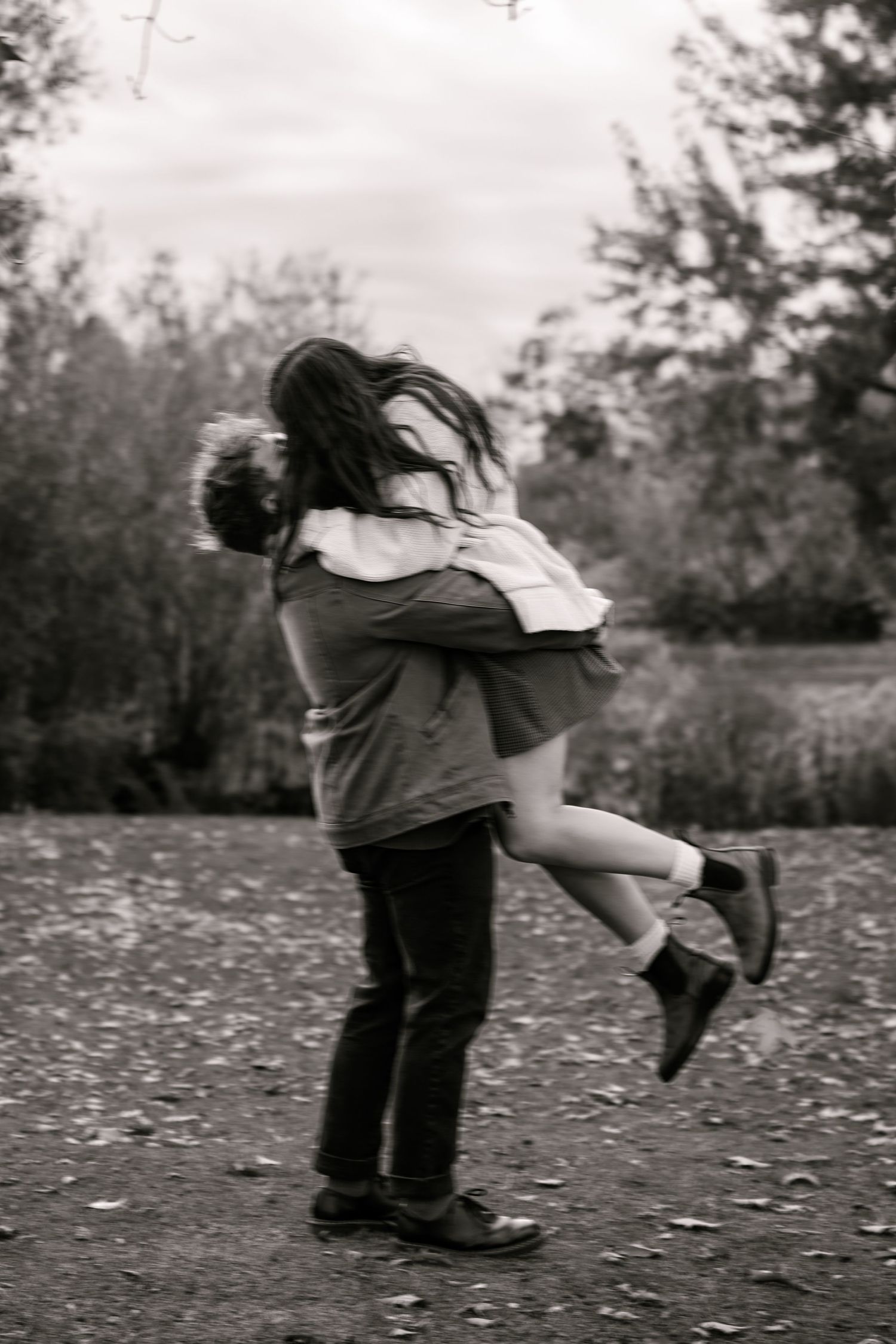 Black and white photo of a couple embracing outdoors, man lifting woman in a romantic park setting.